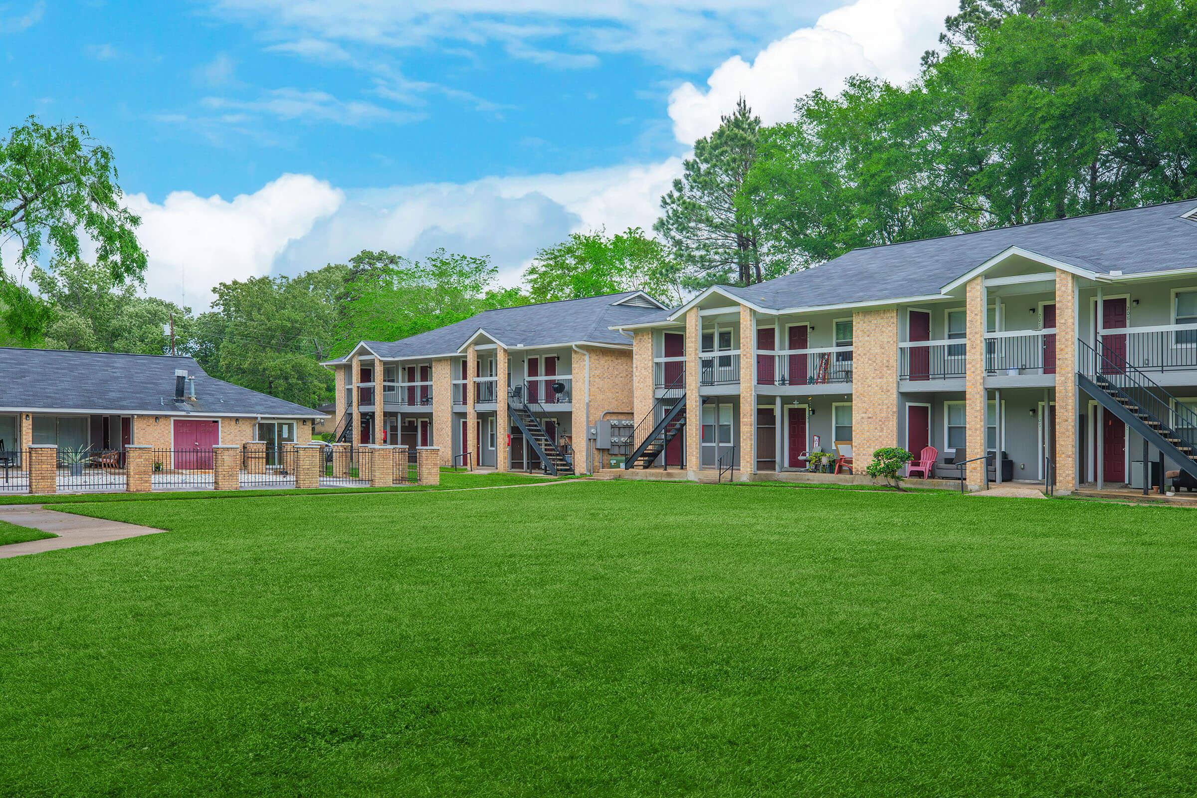 A view of a well-maintained apartment complex featuring two-story buildings with brick facades, balconies, and a vibrant green lawn. Trees and a blue sky with scattered clouds provide a pleasant backdrop, creating a welcoming outdoor environment for residents.