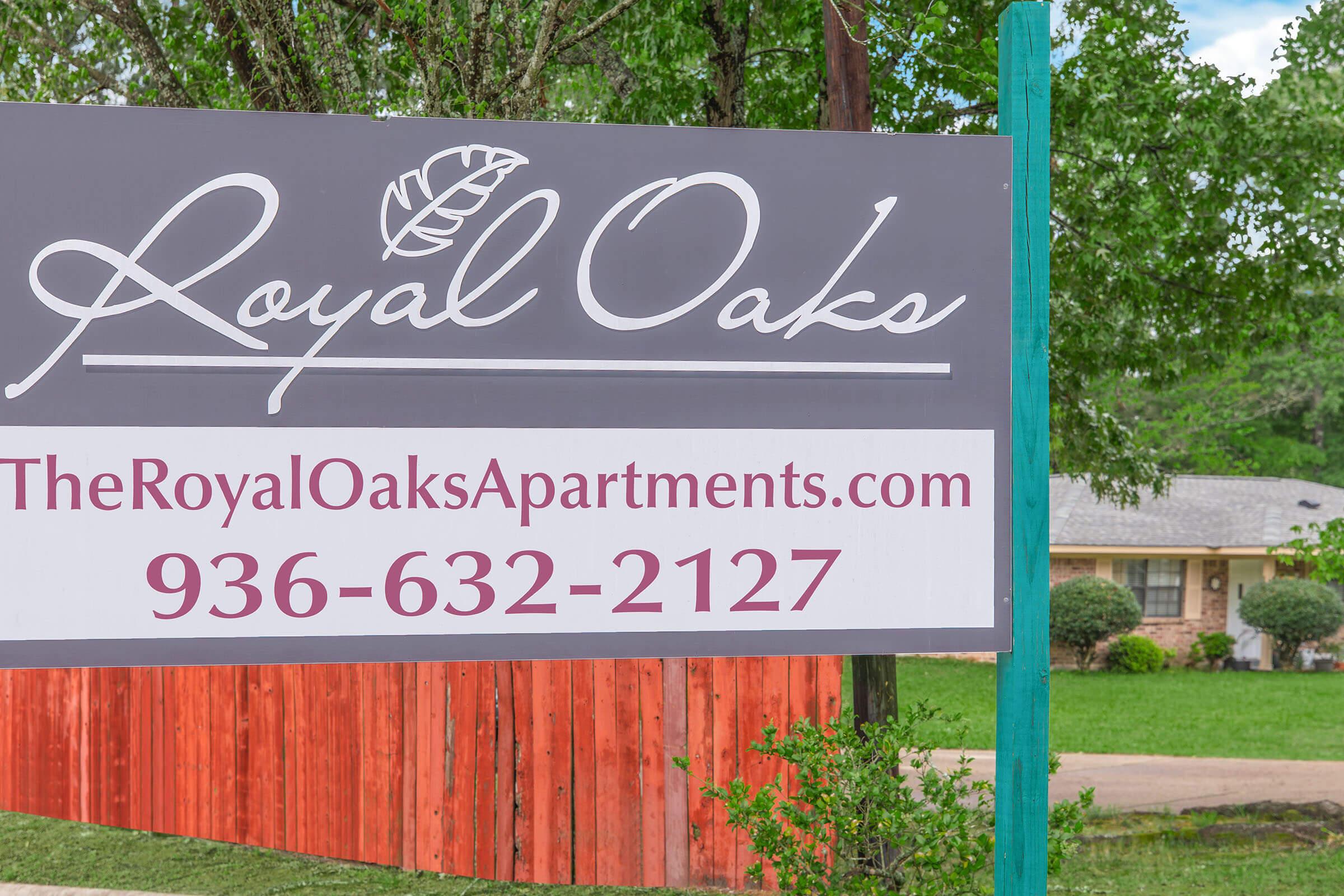 Sign for Royal Oaks Apartments featuring the name, website, and phone number. The sign is supported by a green post, with a background of trees and a red wooden fence visible in the surroundings.