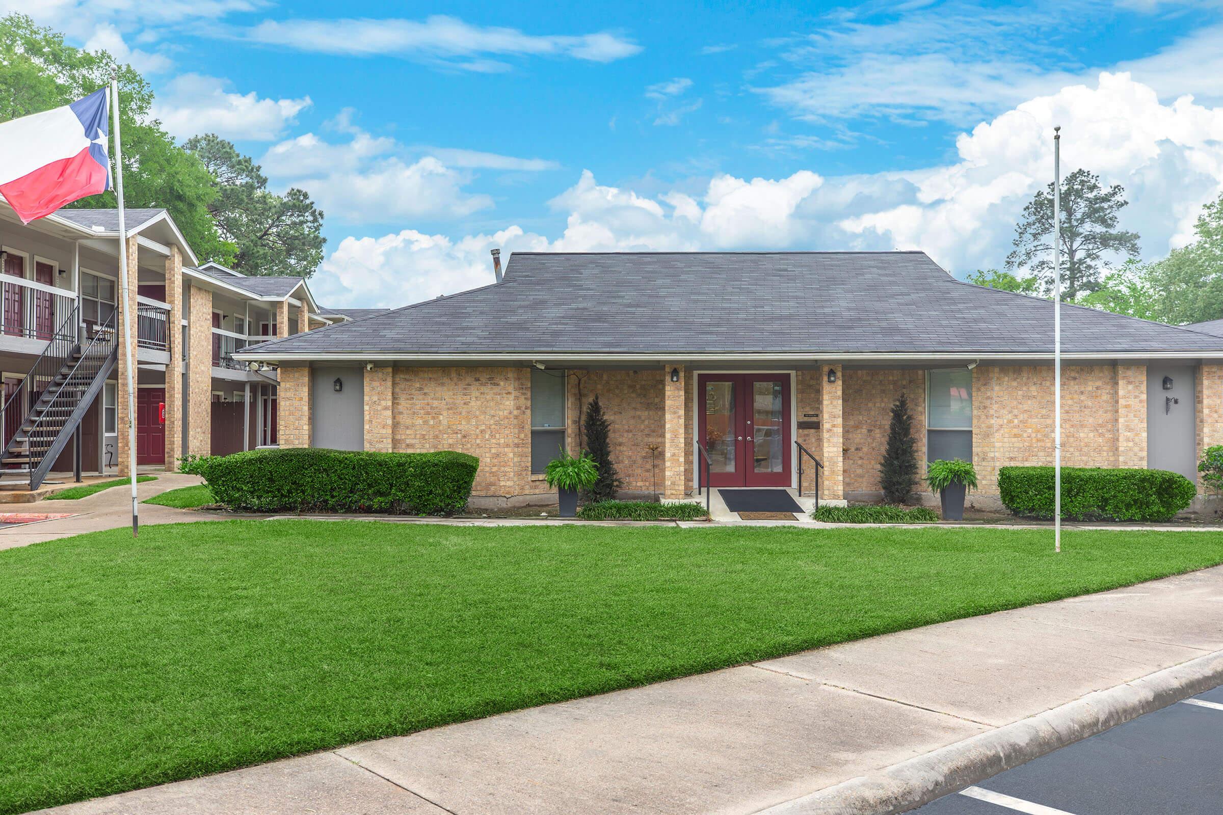 A brick building with a pitched roof, featuring a welcoming entrance with a red door, flanked by two small plants. In the foreground, well-maintained green grass stretches across the lawn. A Texas flag is raised beside the driveway, and neighboring apartment buildings with balconies are visible on the left.