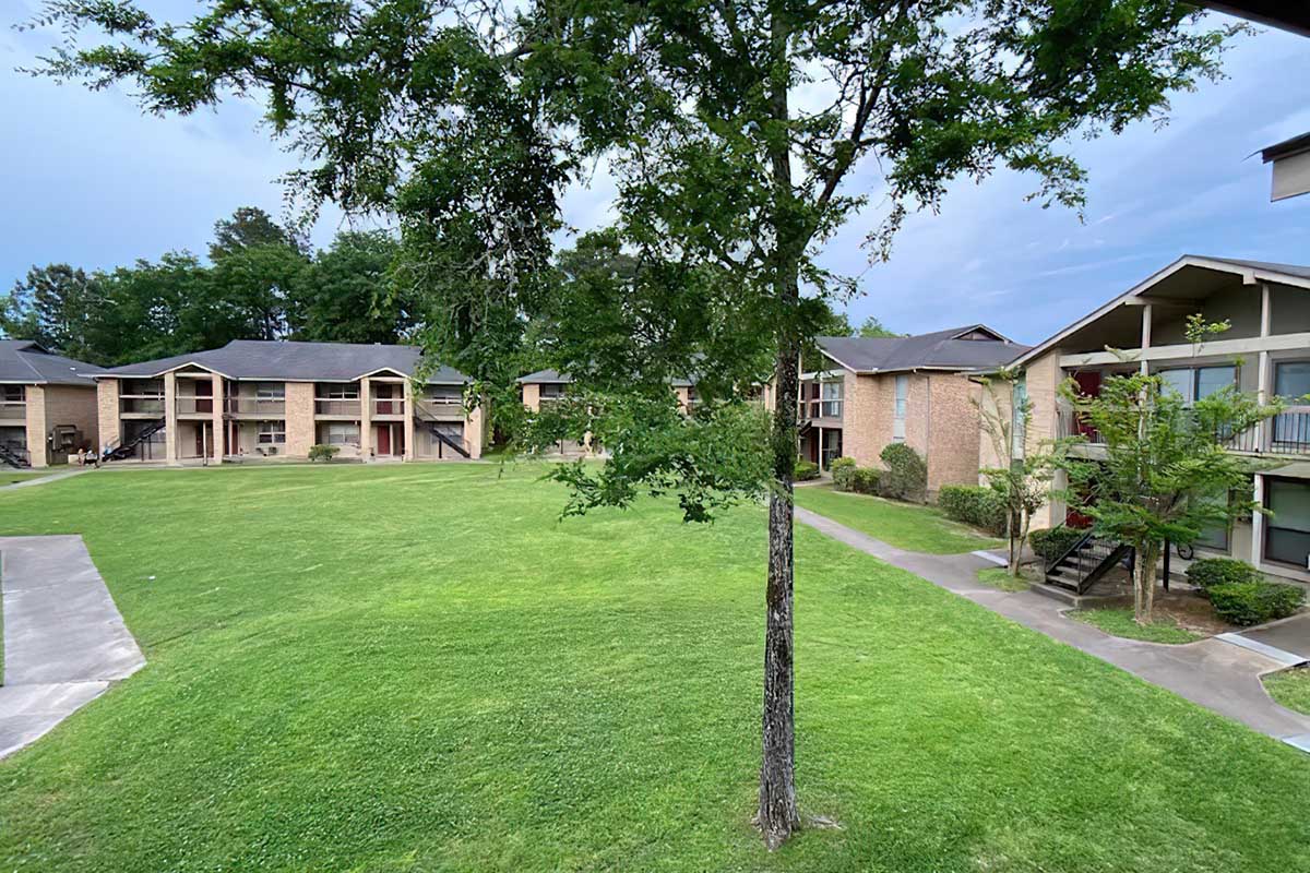A view of a residential complex featuring multiple two-story brick buildings arranged around a grassy courtyard. A tree stands in the foreground, and the overall atmosphere is calm and green, suggesting a suburban setting. The sky is overcast, indicating potential rain.