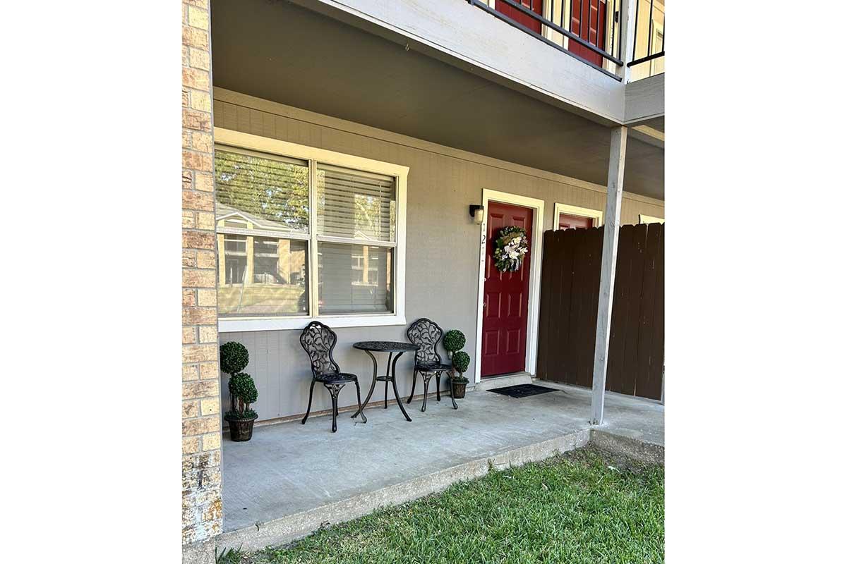 A welcoming front porch with two black metal chairs and a small round table. A decorative wreath is hanging on a red door, and there are potted topiary plants on either side. The porch is surrounded by a brown fence and has a grassy area in front. Sunlight filters in, creating a cozy atmosphere.