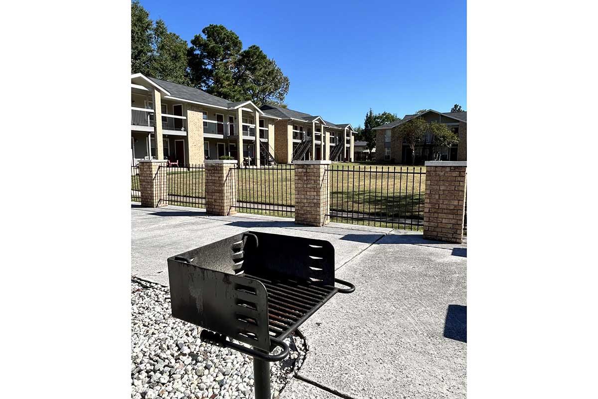 A black charcoal grill on a gravel base, located in a courtyard area. In the background, there are several apartment buildings and a clear blue sky overhead. The scene is well-lit and depicts an outdoor communal space.