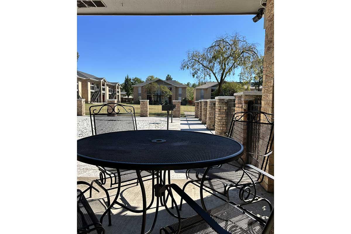 Outdoor scene featuring a black metal dining table surrounded by chairs, viewed from a covered patio. In the background, a clear blue sky and residential buildings are visible, along with a tree and gravel path. The setting appears tranquil and inviting for outdoor gatherings.
