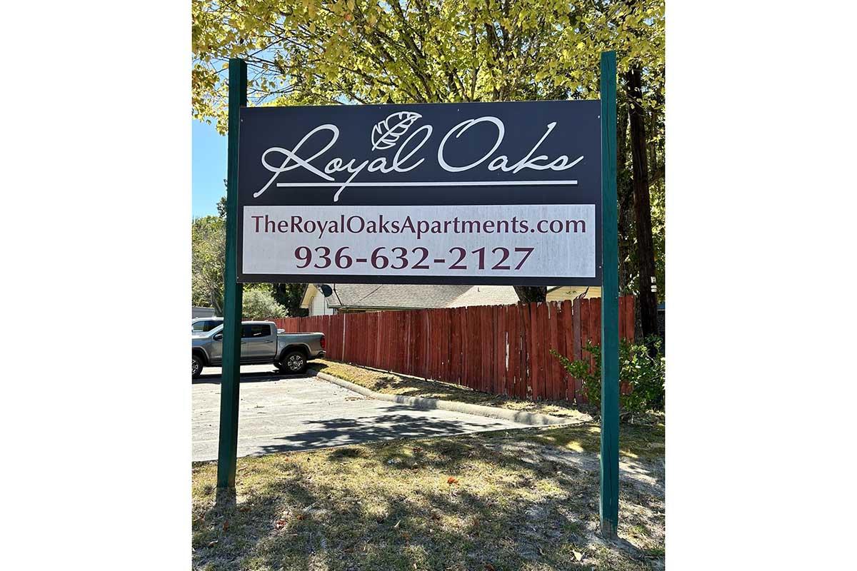 Sign for Royal Oaks Apartments featuring the website TheRoyalOaksApartments.com and phone number 936-632-2127. The sign is positioned outdoors with trees and a red fence visible in the background.