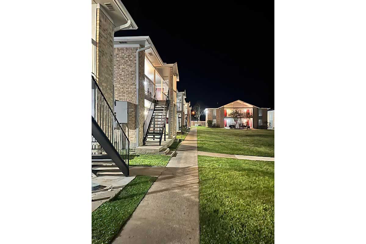 Nighttime view of an apartment complex featuring two-story buildings with brick facades. A well-lit pathway runs between the buildings, leading to a grassy area. Stairs lead to second-floor units, and nearby lights illuminate the surroundings. The scene conveys a calm, residential atmosphere.