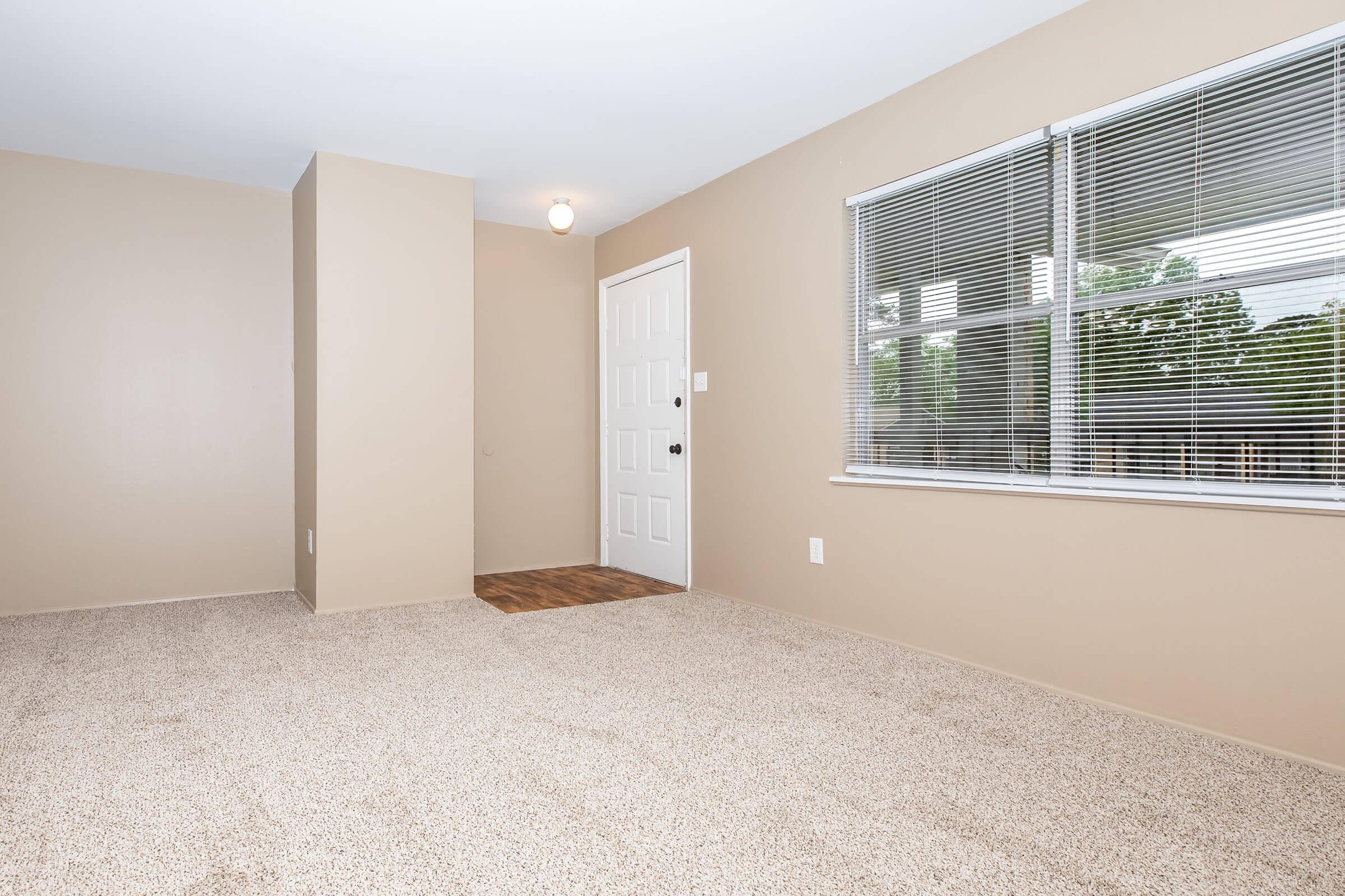 A vacant room with beige walls and carpet, featuring a closed white door and a large window with blinds that lets in natural light. The space is uncluttered, providing a blank canvas for personal decoration. The flooring near the entrance appears to be wood.