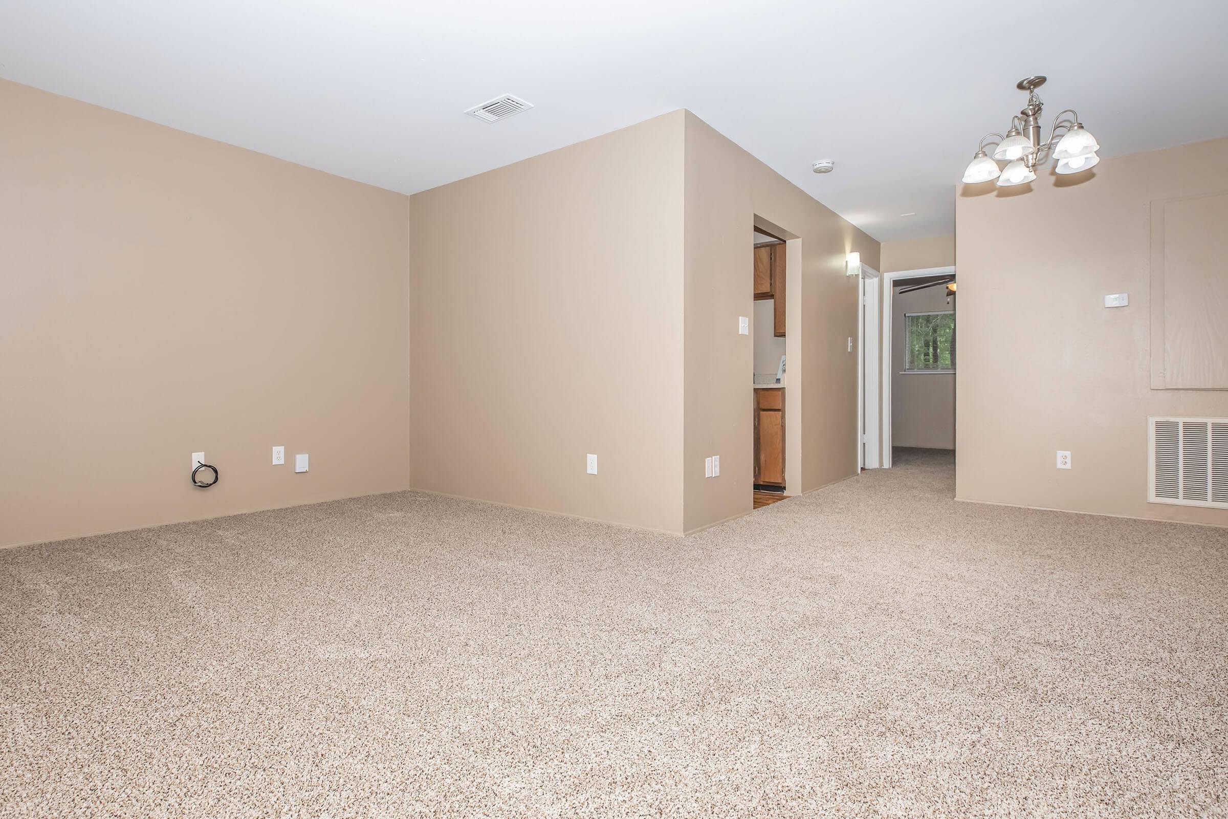 A spacious, empty room with light beige walls and plush carpet flooring. The area features a doorway leading to a kitchen, and a light fixture with three bulbs hanging from the ceiling. There are no furniture items present, creating a clean and airy atmosphere.