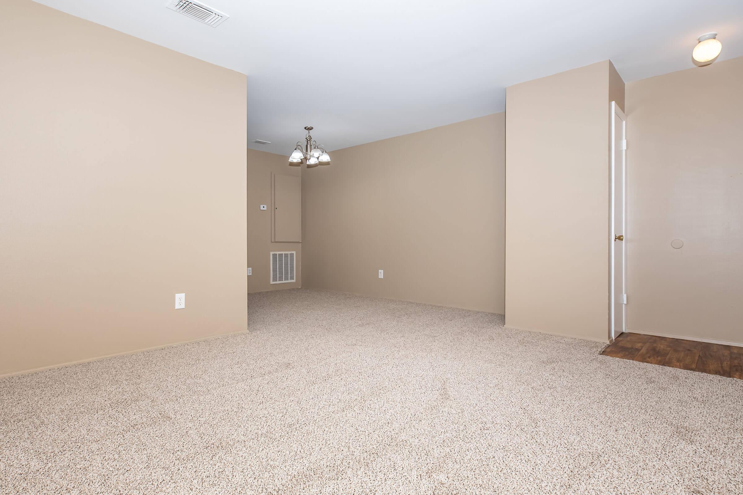 A spacious, empty room with light beige walls and carpet. A small chandelier hangs from the ceiling, illuminating the area. To the right, there is a doorway leading out of the room. The overall appearance is clean and simple, providing a blank canvas for future decoration or furniture placement.