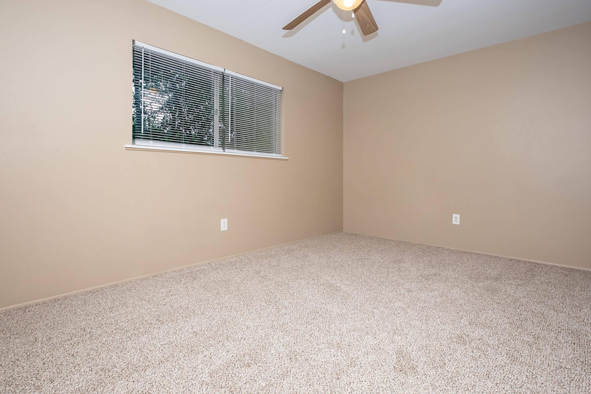 A spacious, empty room with beige walls and light brown carpet. There is a window with blinds on the left side, allowing natural light to enter. A ceiling fan is visible, and there are no furnishings or decorations, giving the room a minimalist feel.