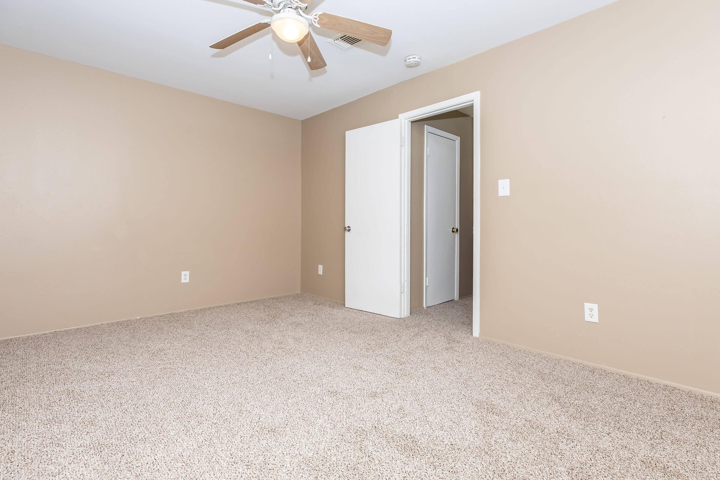 A spacious, empty room with light beige walls and a light brown carpet. There is a ceiling fan overhead and a closed door on the right leading to another room. The room is well-lit, showcasing its clean and simple design.