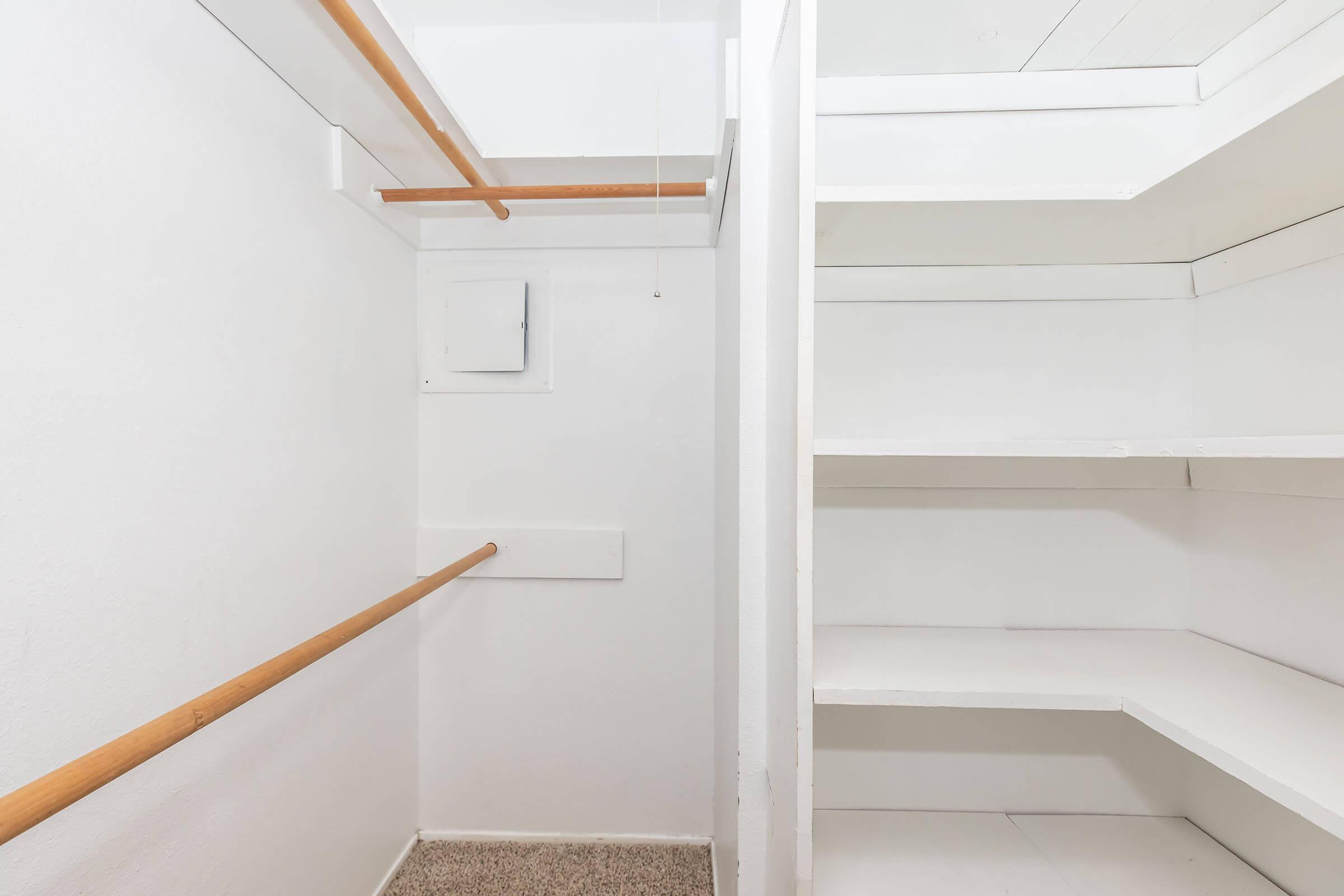 An empty closet with white walls and shelving. On the left, there is a wooden rod for hanging clothes. The floor is carpeted and there is a small panel on the wall. The overall space is bright and uncluttered, providing a minimalist storage area.