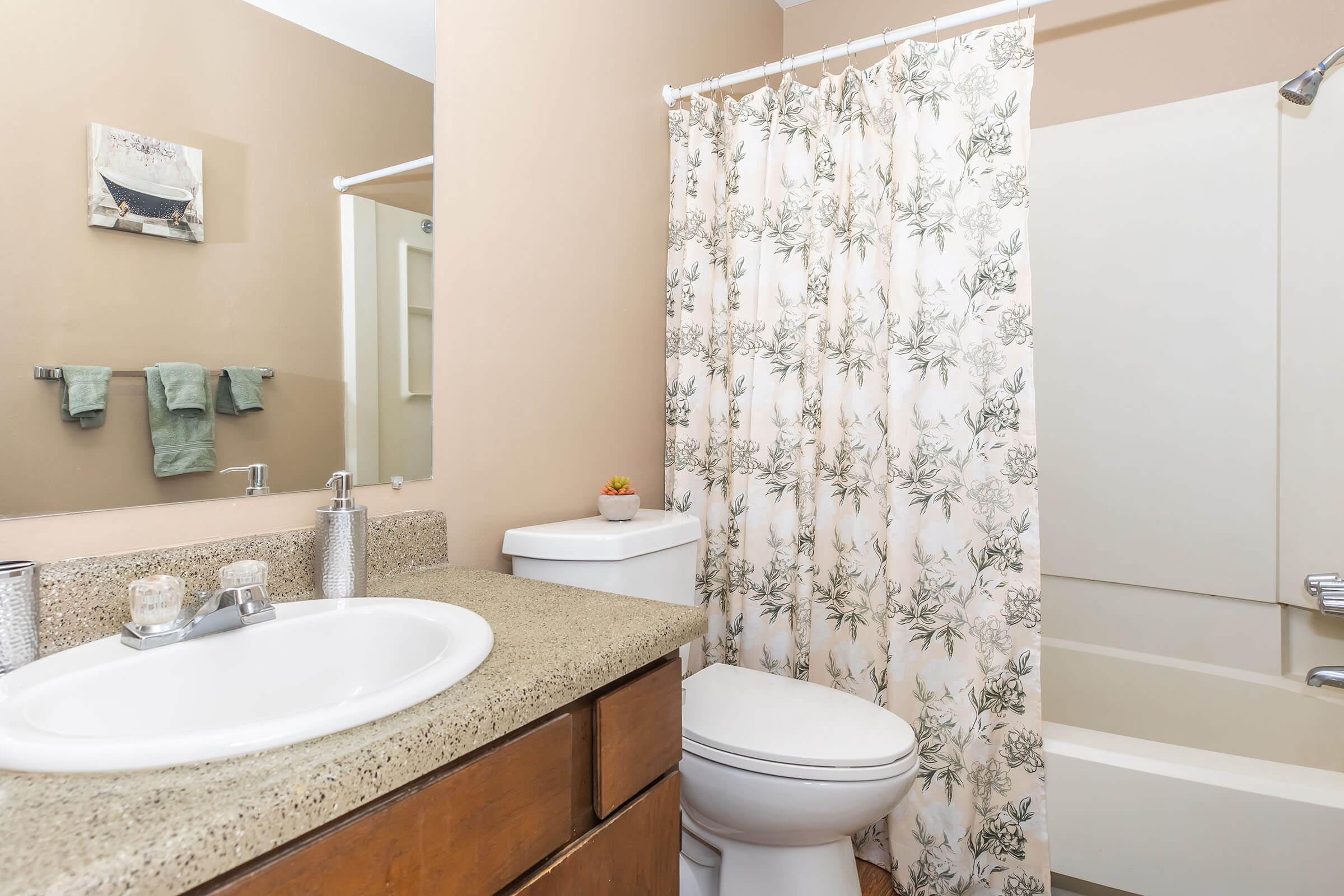 A clean and tidy bathroom featuring a light brown color scheme. It includes a round sink with a granite countertop, a toilet, and a bathtub with a shower curtain displaying a floral pattern. There is a mirror above the sink and some green towels hanging on a rack. A small decorative plant adds a touch of greenery.