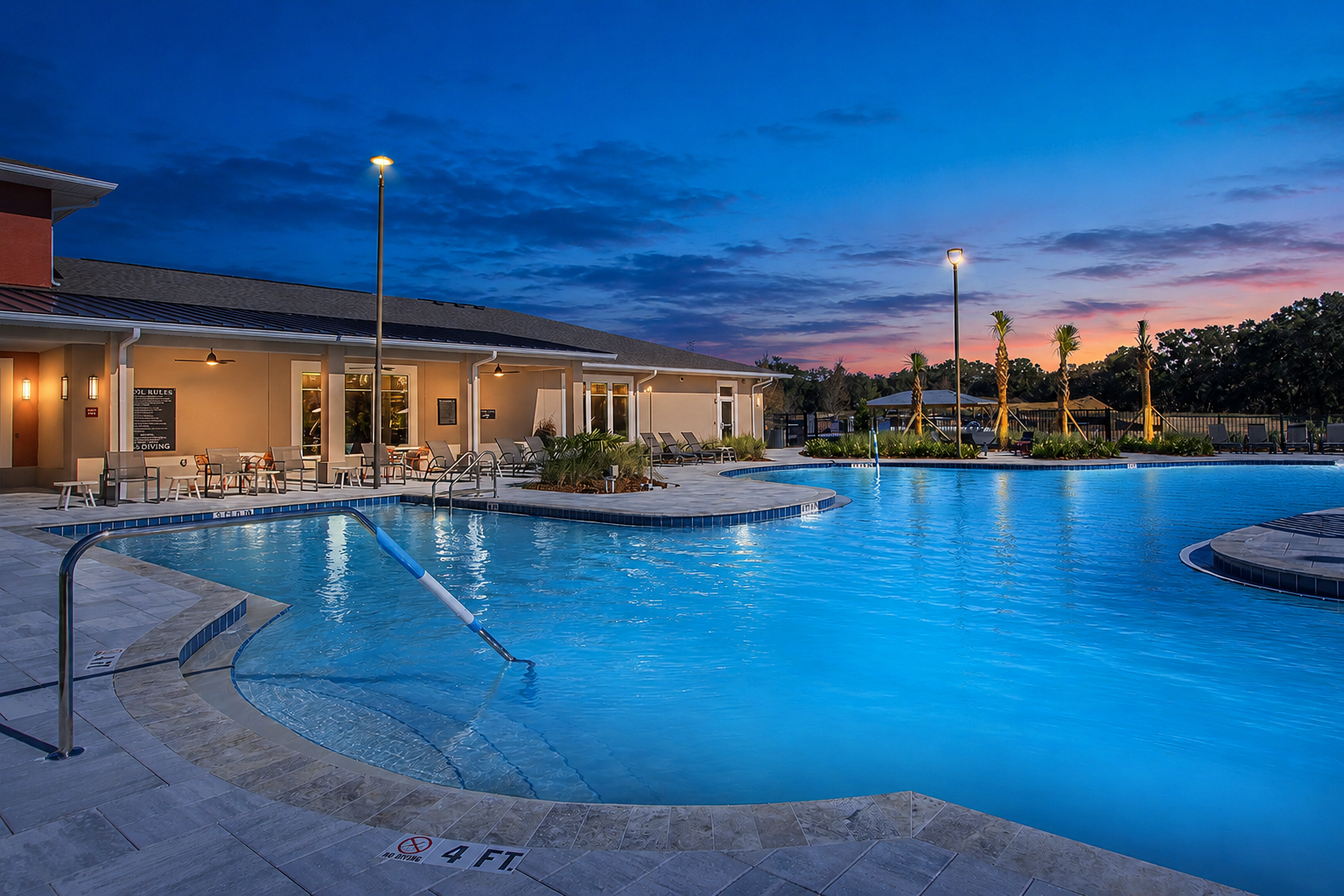 A scenic view of a swimming pool at dusk, featuring soft blue water illuminated by lights. Surrounding the pool are lounge chairs and palm trees, with a building visible in the background. The sky showcases beautiful shades of blue and orange, creating a tranquil atmosphere.