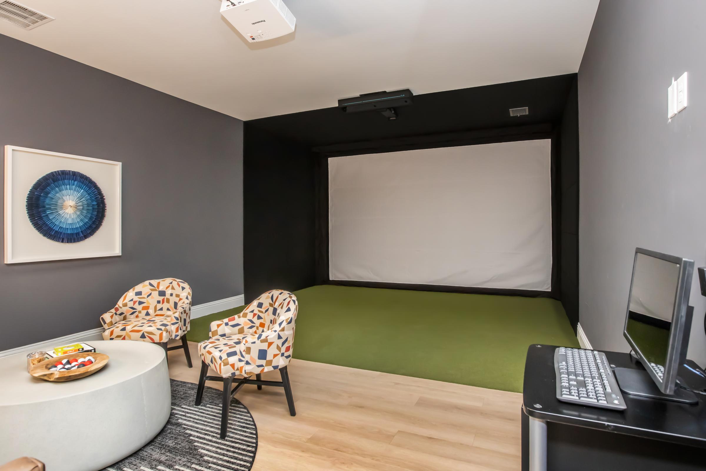A cozy room featuring two patterned chairs and a modern round coffee table, with a large blank projection screen against a dark wall. A small monitor and keyboard are on a black desk, while the floor has a green carpet. Wall art is displayed nearby, adding a decorative touch to the space.