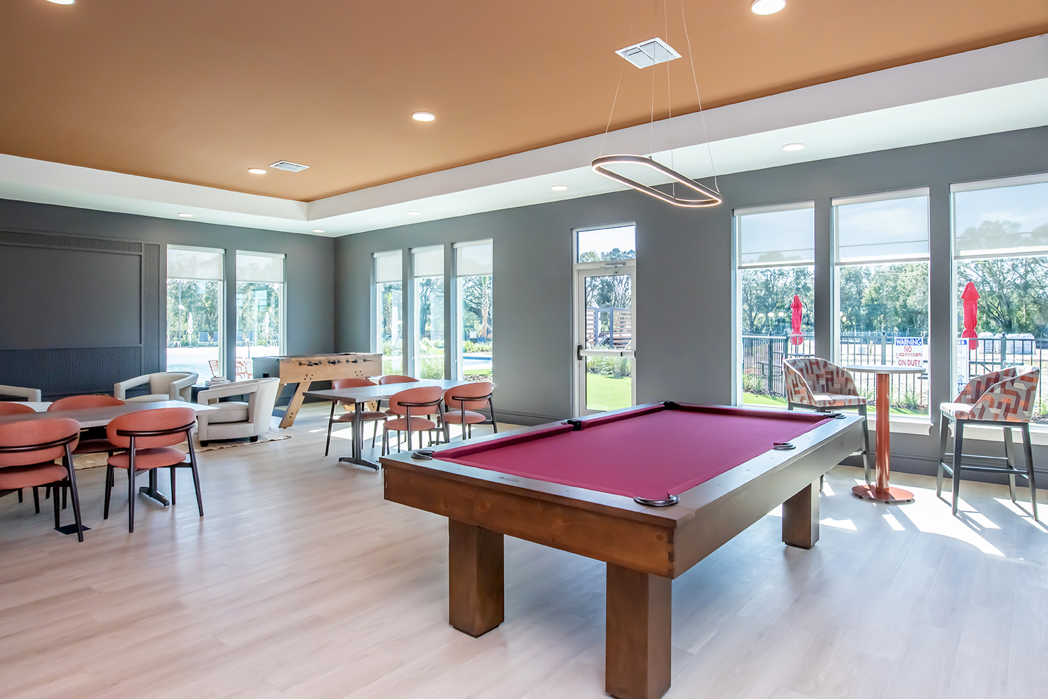 A bright, modern game room featuring a pool table with a burgundy felt top, surrounded by seating areas. Large windows provide natural light and a view of the outdoors, with contemporary furniture and decor creating a stylish atmosphere.