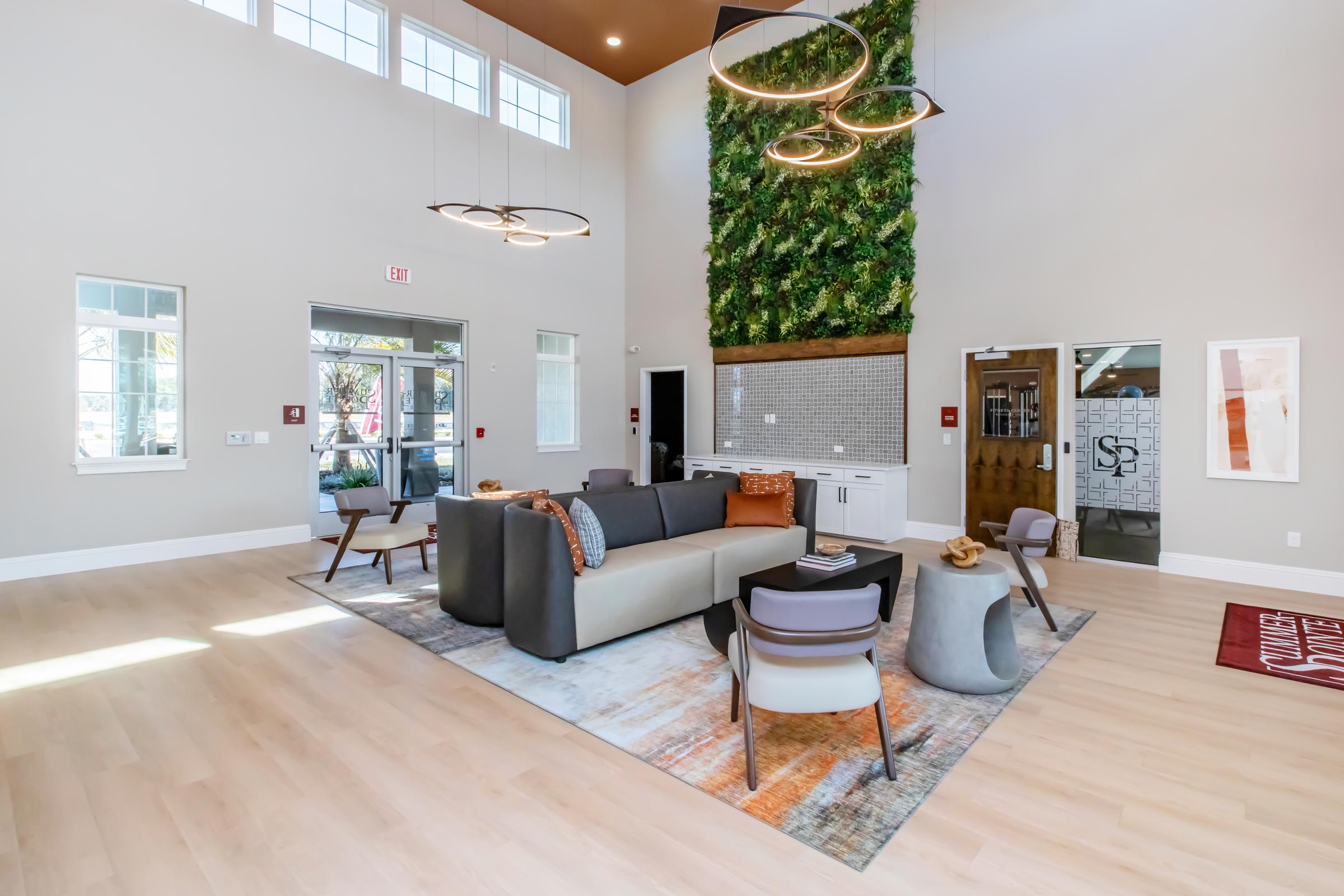 A spacious and modern common area featuring high ceilings, large windows, and a vertical garden wall. There are comfortable seating options including a gray sofa, decorative chairs, and a coffee table on a patterned rug. The room is well-lit and has stylish lighting fixtures, creating a welcoming atmosphere.