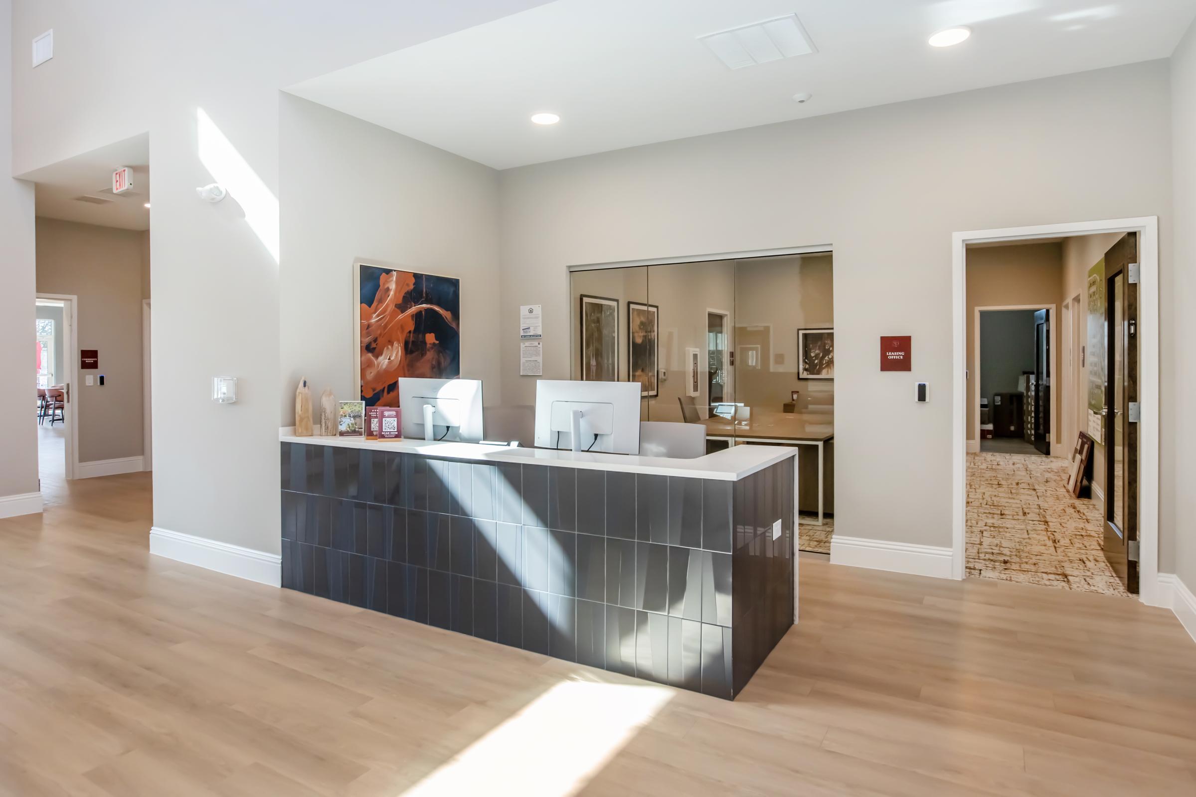 Modern reception area with a sleek front desk featuring two computer monitors. The walls are painted light gray, and there's artwork on the left. Sunlight streams through windows, illuminating the space, which has wooden flooring and paths leading to adjacent rooms.