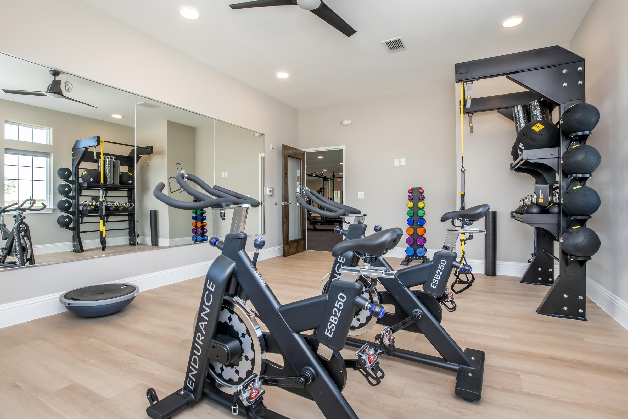A modern home gym featuring multiple stationary bikes, a weight training station with various weights, a large mirror, and hardwood flooring. The room is well-lit, with a ceiling fan, creating an inviting workout environment.