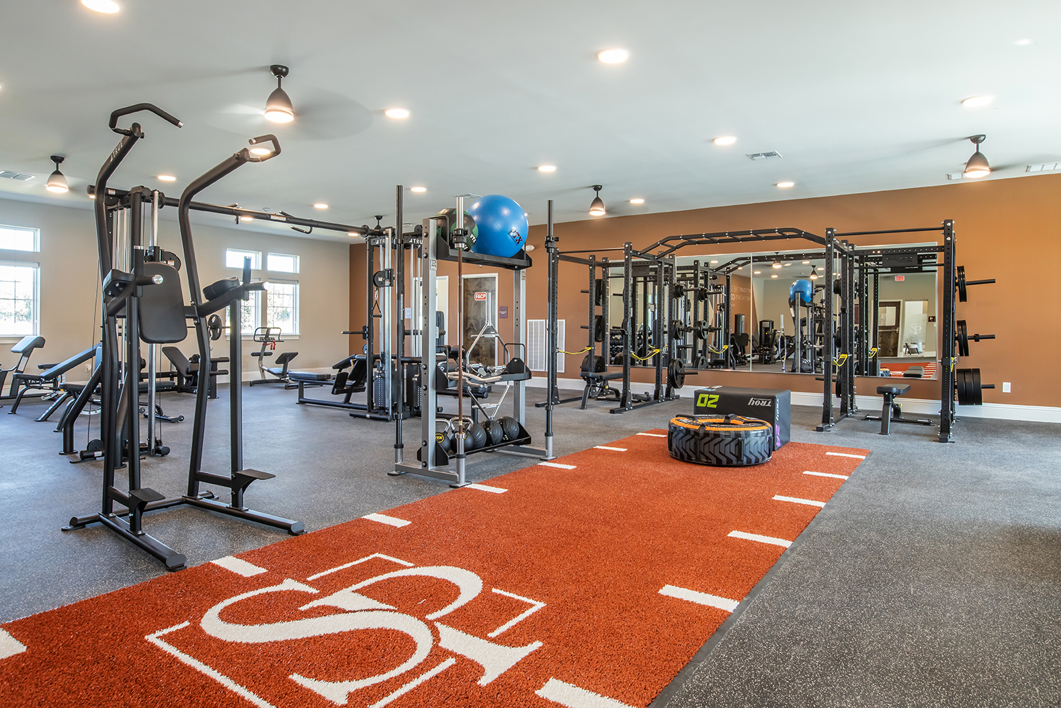 Modern gym interior featuring fitness equipment such as weight machines, free weights, exercise mats, and a large tire for workouts. The flooring has a red and black design with a logo, well-lit with multiple ceiling lights and large windows. The space is clean and well-organized, promoting an active lifestyle.