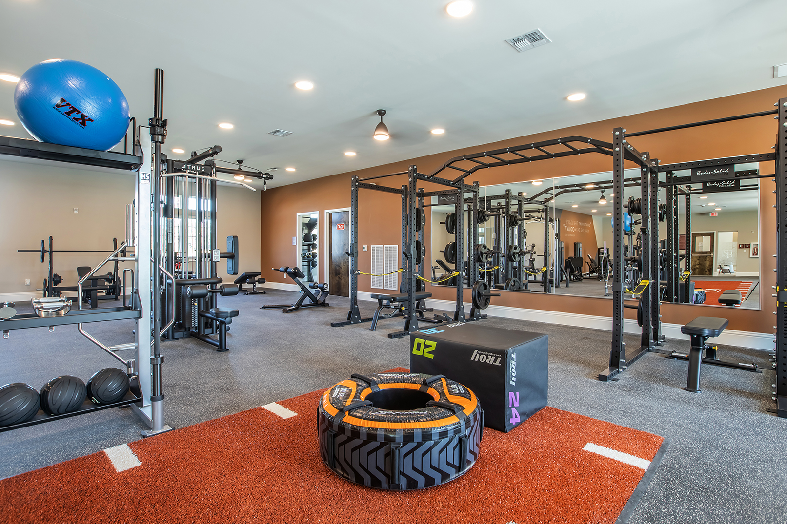 A modern gym interior featuring various workout equipment. There are free weights, a fitness ball, a weight bench, a squat rack with pull-up bars, and a rubber tire on a turf area. Mirrors reflect the space, and the walls are painted in warm tones, enhancing the inviting atmosphere for fitness training.