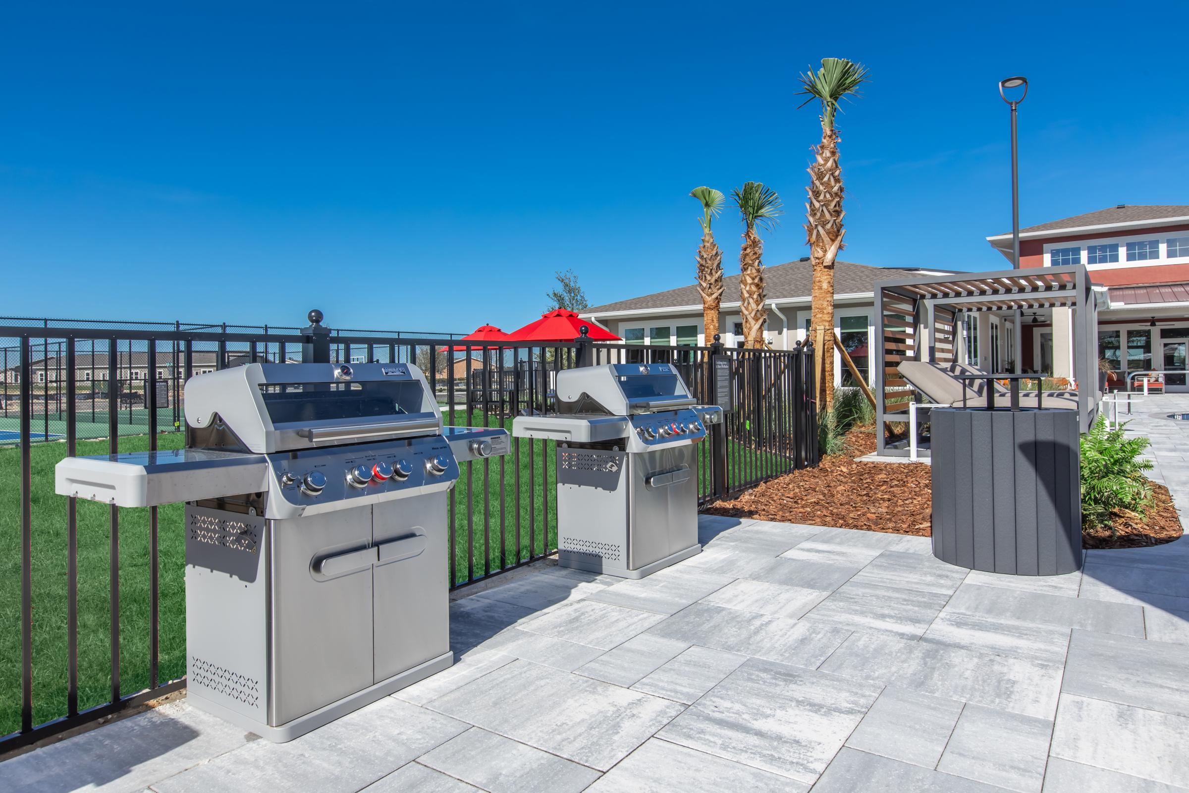 Outdoor grilling area featuring two stainless steel gas grills surrounded by a black fence. Palm trees are visible in the background, along with a pool and a modern building. The patio has gray stone tiles and is well-lit under a clear blue sky, creating an inviting space for barbecues and gatherings.