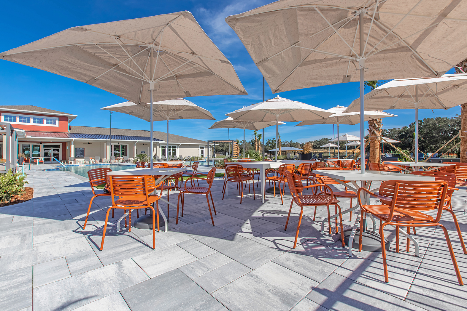 Outdoor dining area featuring orange metal chairs and large umbrellas on a patio with gray stone flooring. In the background, a building with large windows and palm trees is visible under a clear blue sky.
