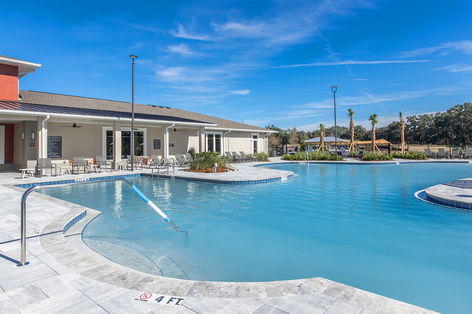 A sparkling blue pool surrounded by a modern building. The pool features a shallow area with a depth indicator and is lined with lounge chairs. In the background, landscaped greenery and play structures can be seen under a clear blue sky.