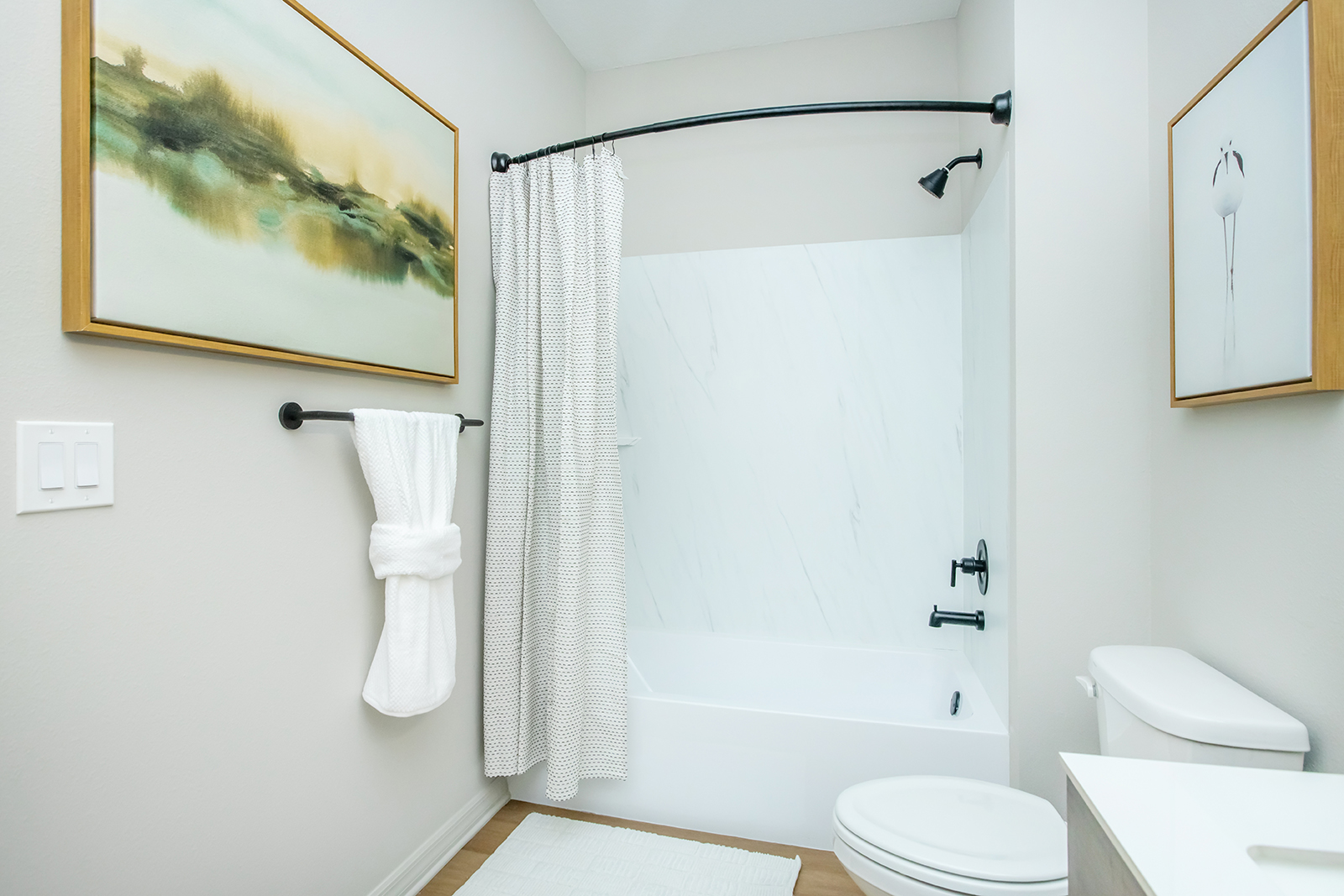 A modern bathroom featuring a bathtub with a shower curtain, a white toilet, and a sink with a cabinet. There are two framed artworks on the walls, one depicting a landscape and the other a floral theme. A towel is hung on a towel rack, and the floor has a textured white rug.