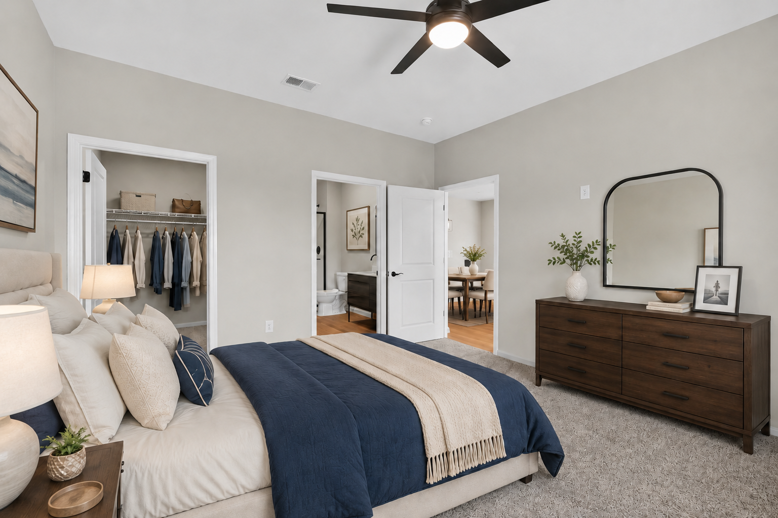 A modern bedroom featuring a king-sized bed with blue and beige bedding, accent pillows, and a beige throw. The room has light gray walls, a ceiling fan, and a wooden dresser with decorative items. A mirror hangs above the dresser, and two doors lead to a closet and another room. Soft lighting creates a cozy atmosphere.