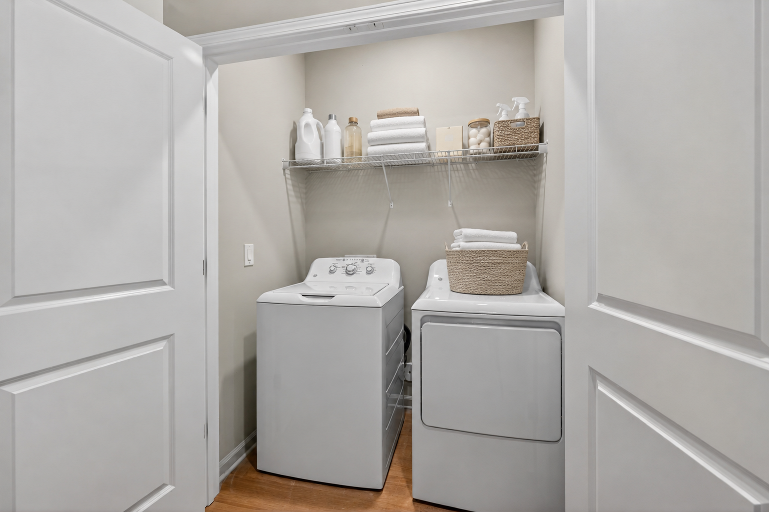 A small laundry room featuring a washing machine and dryer side by side. Above, a wire shelf holds neatly stacked towels and various cleaning supplies. The walls are painted a light color, and the floor has wooden planks. Two white doors open into the space.