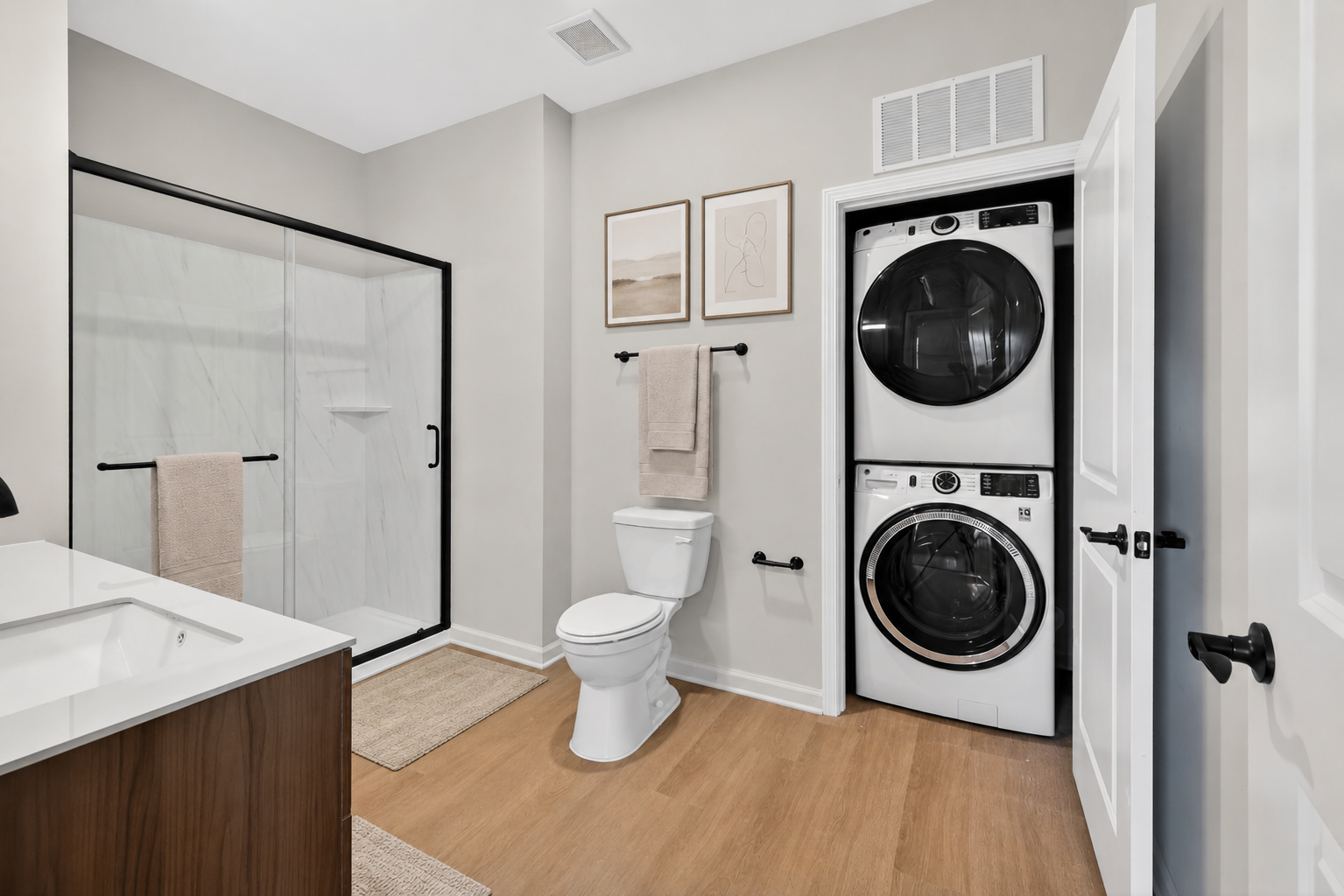 A modern bathroom featuring a glass shower, a toilet, a double washer and dryer stacked in a closet, and minimalist decor including two framed pictures on the wall. The color scheme is light, with beige and white tones and wood accents on the floor and cabinetry.