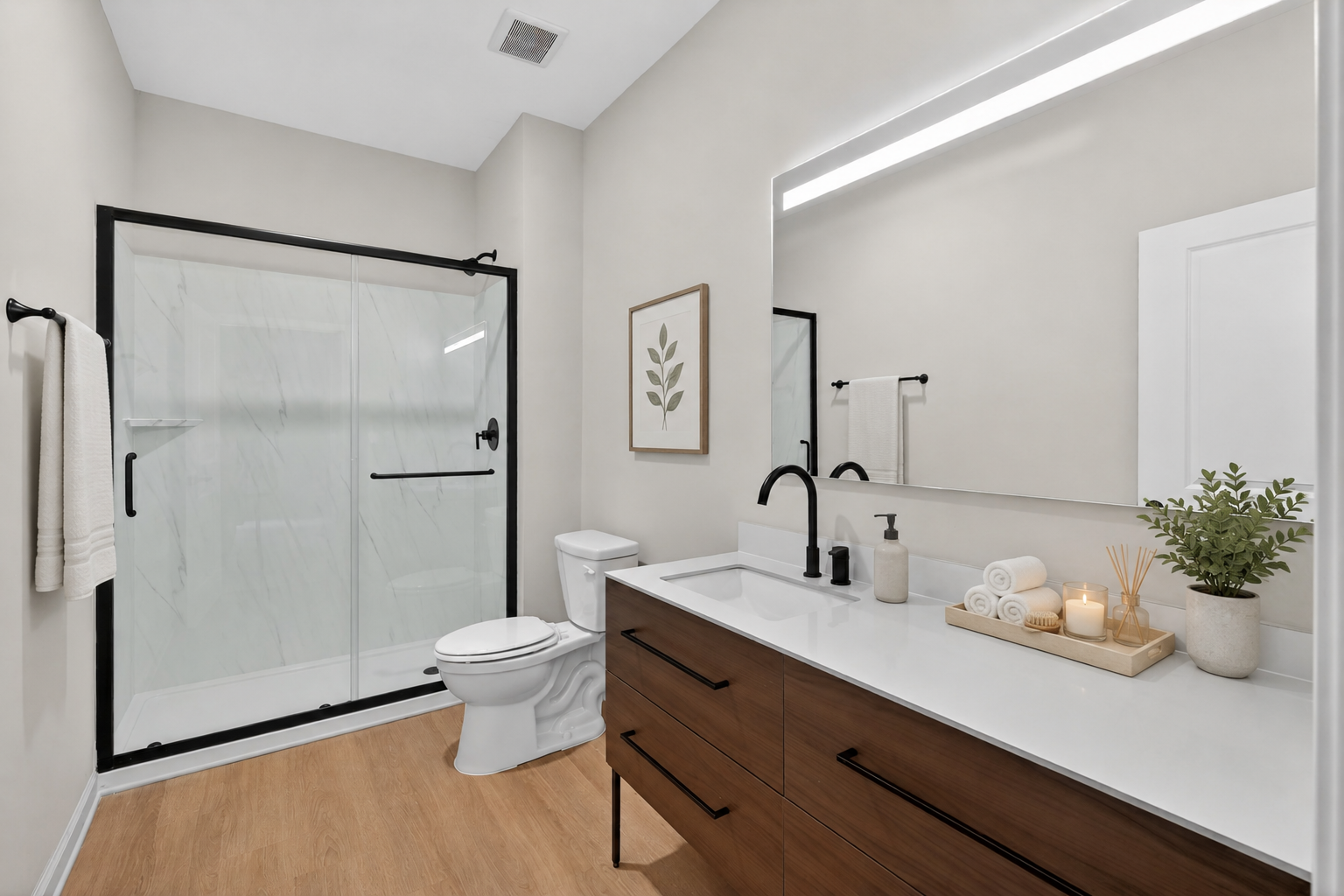 Modern bathroom featuring a walk-in shower with glass doors, a sleek vanity with dark wood drawers, a large mirror with LED lighting, a white toilet, and decorative elements like a potted plant and neatly arranged towels and candles. Walls are painted in a neutral tone, and the flooring is light wood.