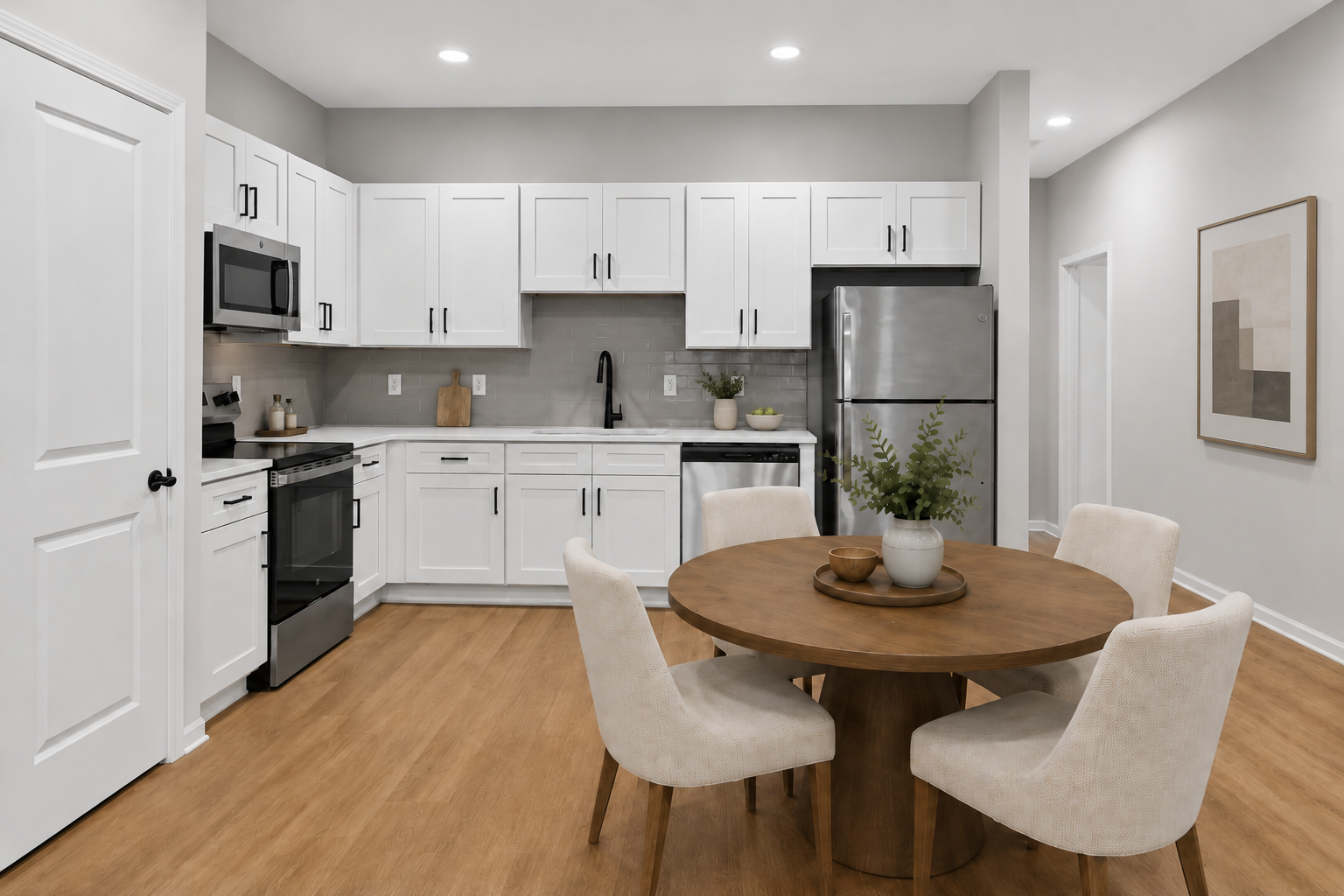 Modern kitchen featuring white cabinetry, stainless steel appliances, and a gray tile backsplash. A round wooden dining table is surrounded by four upholstered chairs. The space is well-lit with recessed lighting, and a potted plant adds a touch of greenery. Neutral colors create a contemporary, inviting atmosphere.