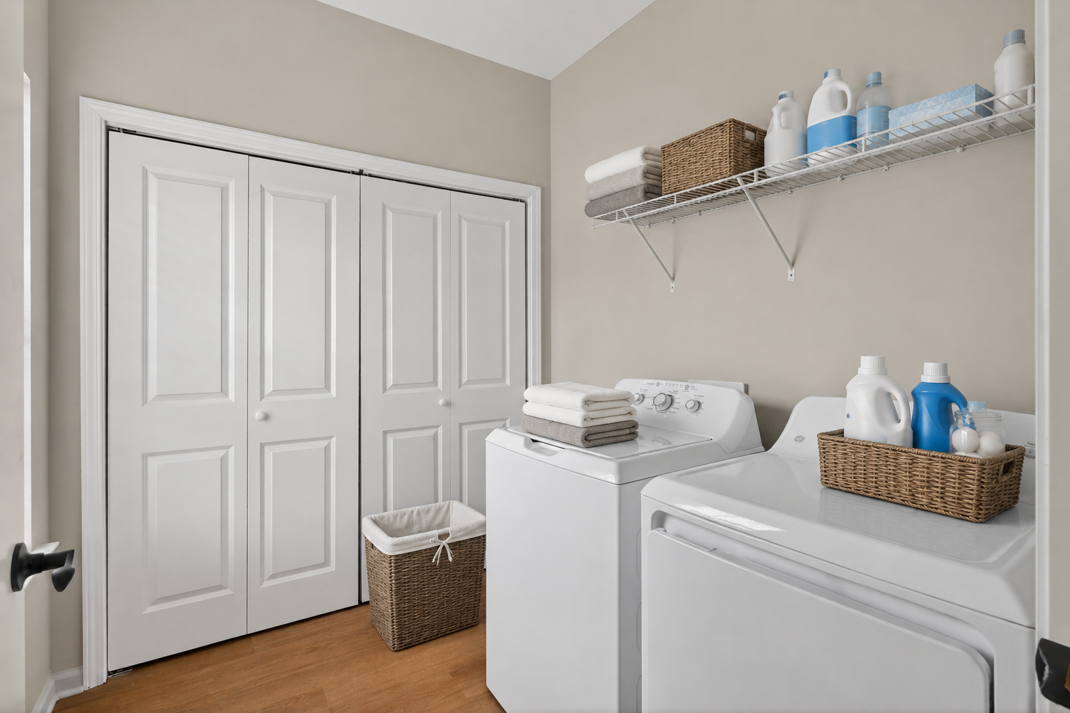 A clean and organized laundry room featuring a washer and dryer stacked next to a shelf with various laundry detergents. A basket holds folded towels, and there's a wicker hamper for dirty clothes. The walls are a soft beige, and there are white double doors leading to a closet.