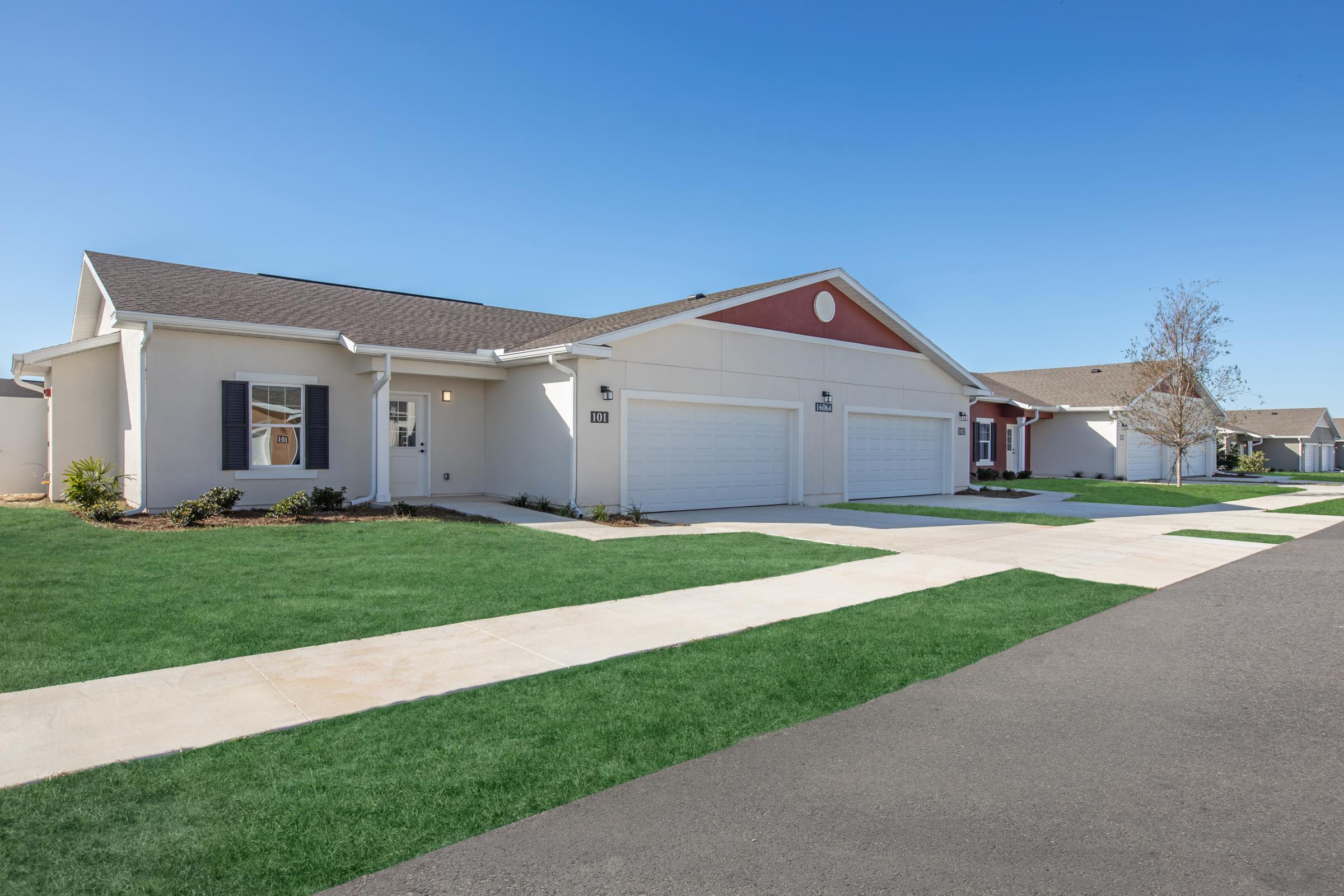 View of a row of modern single-family homes with garages, surrounded by well-maintained lawns and sidewalks. The clear blue sky enhances the appearance of the neighborhood, showcasing a neat and orderly residential area.
