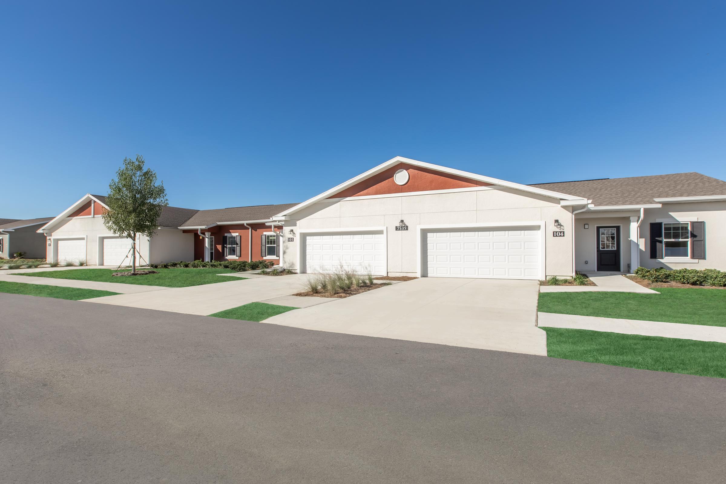 A suburban residential street featuring several modern homes with attached garages. Each house has a well-maintained lawn and clear blue skies overhead. The clean sidewalk and neatly landscaped gardens contribute to a tidy neighborhood atmosphere.