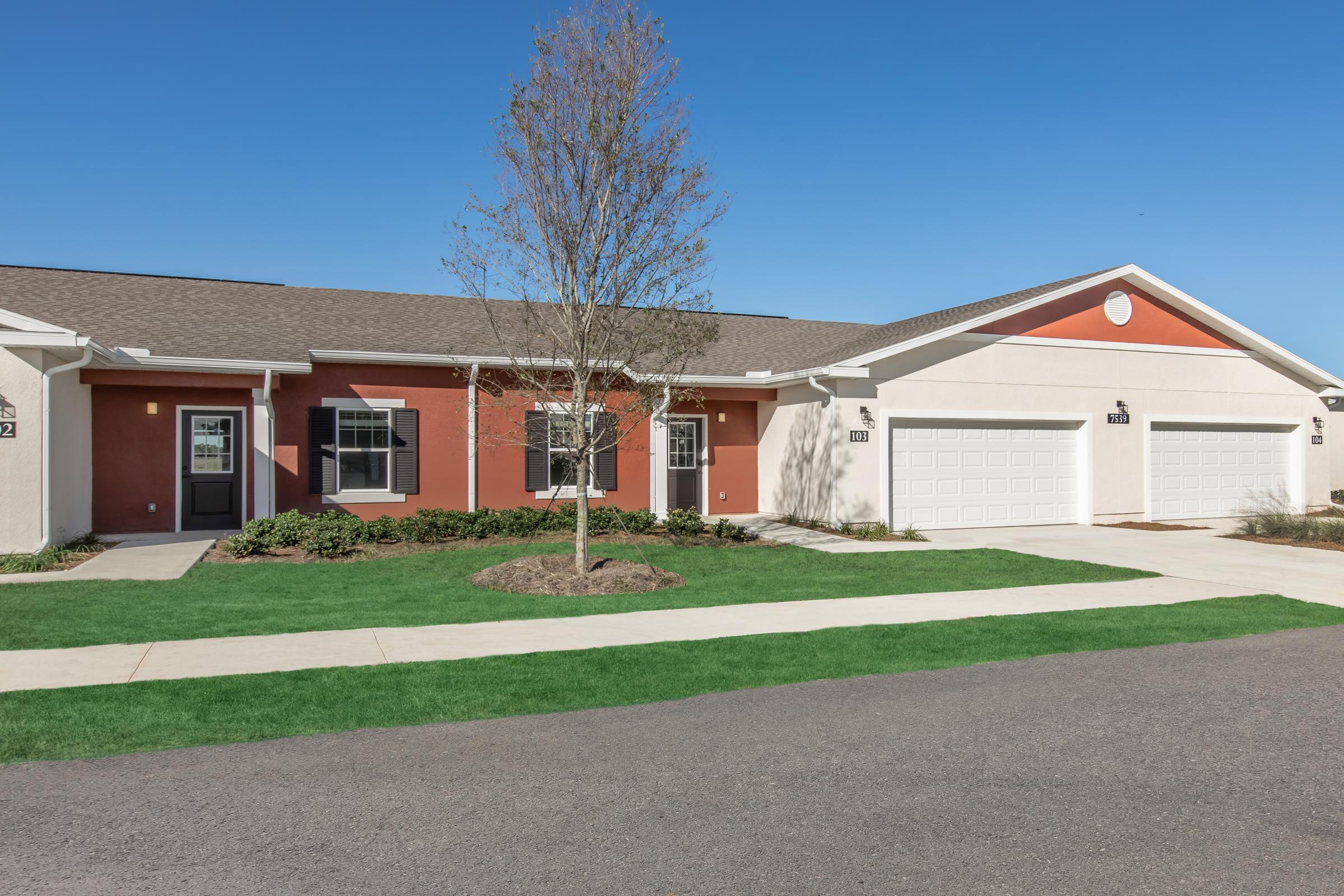A modern single-story residential building featuring a mix of tan and red exterior walls. It has a well-maintained lawn with a small tree in front, clear blue skies overhead, and two garages with white doors visible. The pathway leads up to the entrance, highlighting a welcoming atmosphere.