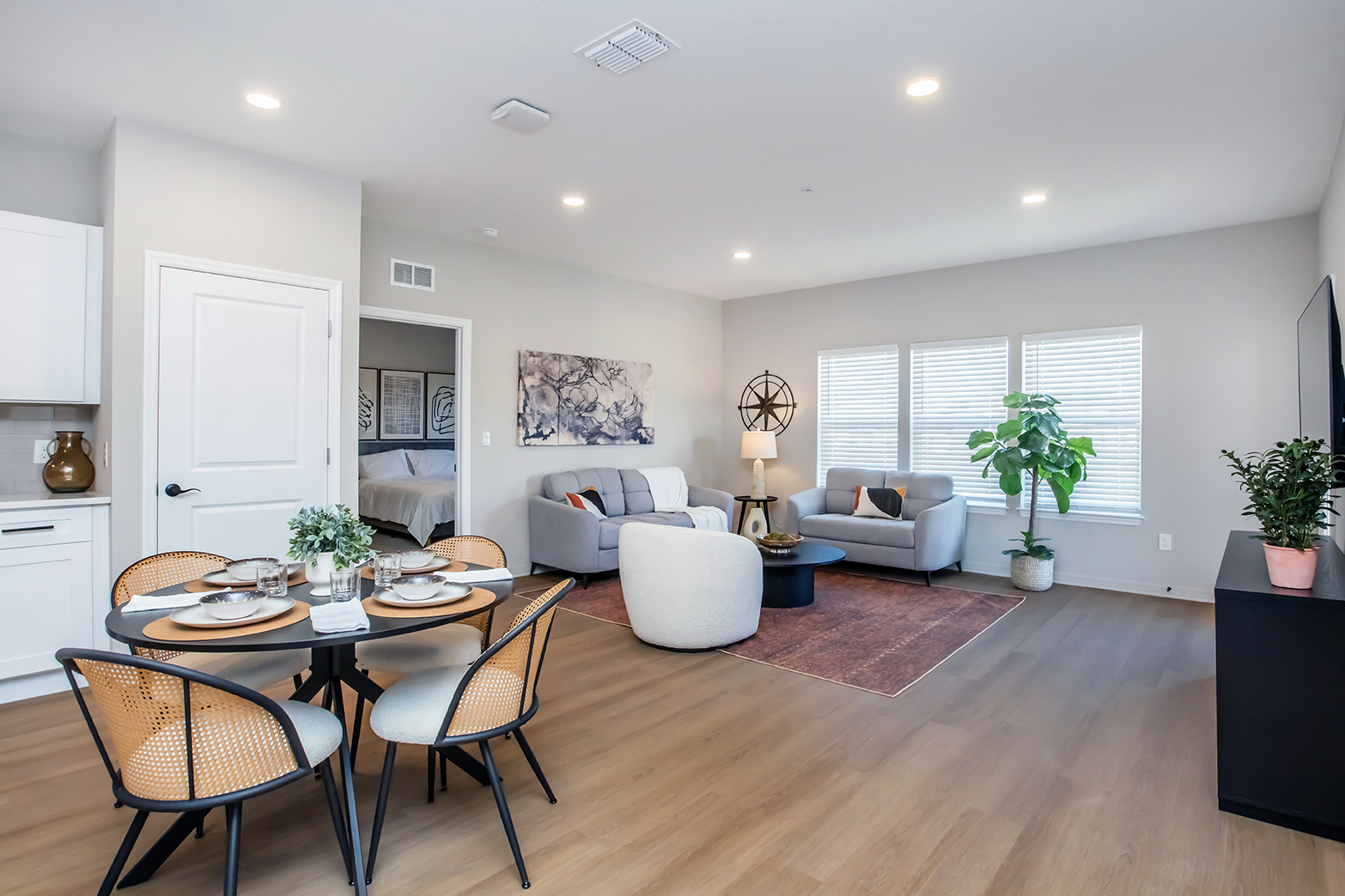 Modern, open-concept living space featuring a dining table set for four, two gray sofas, a stylish coffee table, and a large TV unit. Soft natural light streams through three windows. The decor includes a plant and abstract wall art, creating a cozy and inviting atmosphere.
