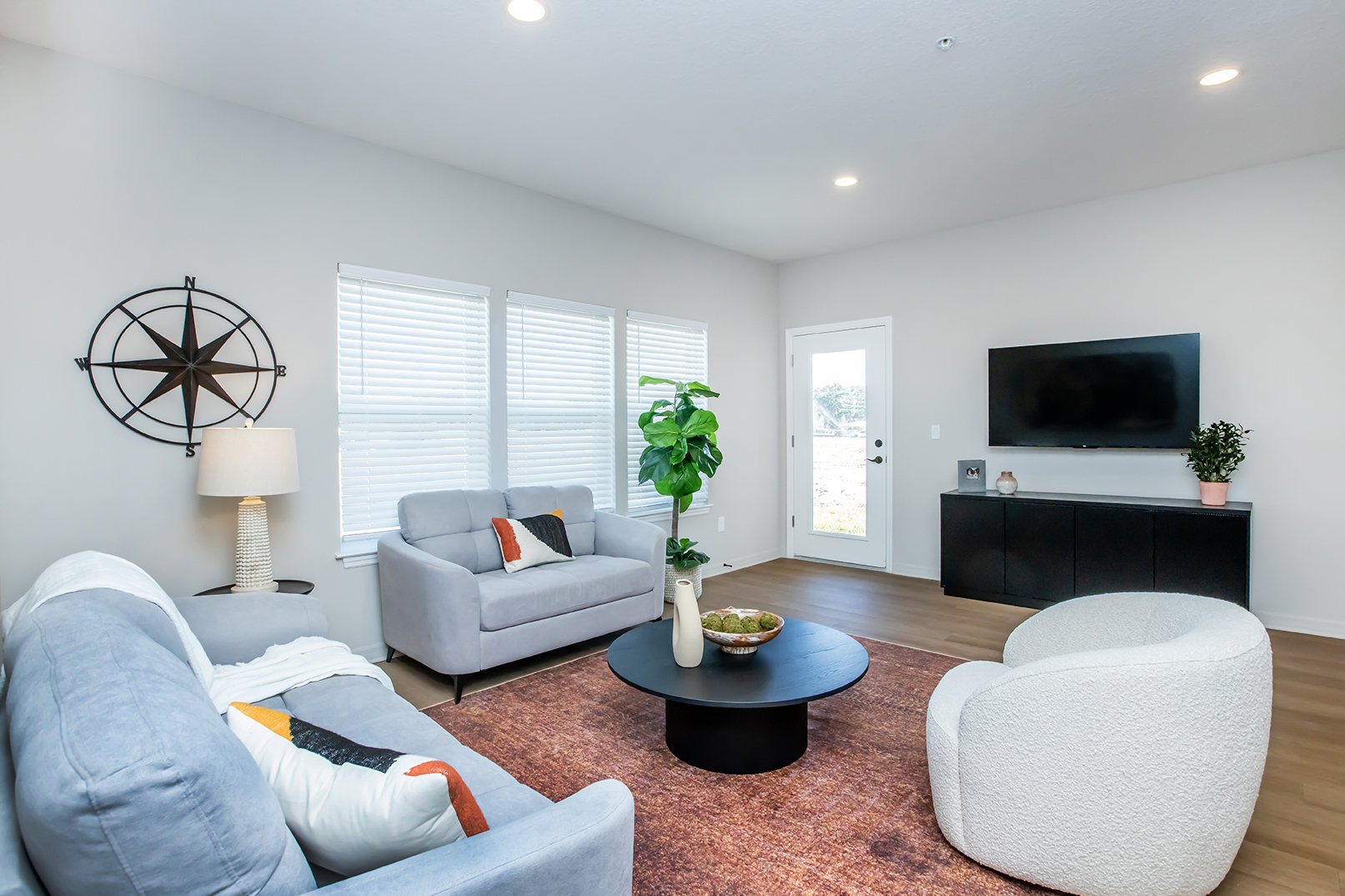 A modern living room featuring two stylish sofas, a round black coffee table, and a decorative bowl. There’s a large television on a dark cabinet, a potted plant beside it, and a star-shaped wall decor. Sunlight filters through the windows, highlighting the minimalist, cozy atmosphere.