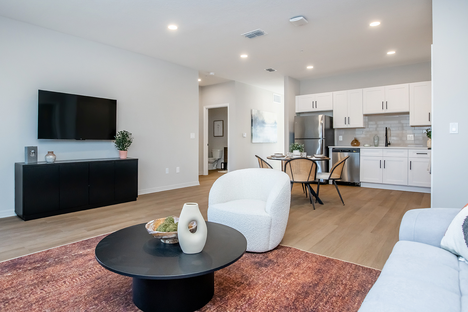 Modern living room and kitchen area featuring a large flat-screen TV, a cozy white chair, a round black coffee table with decor, and a stylish dining table with chairs. The kitchen showcases white cabinetry and stainless steel appliances, all set against light-colored walls and warm wood flooring.
