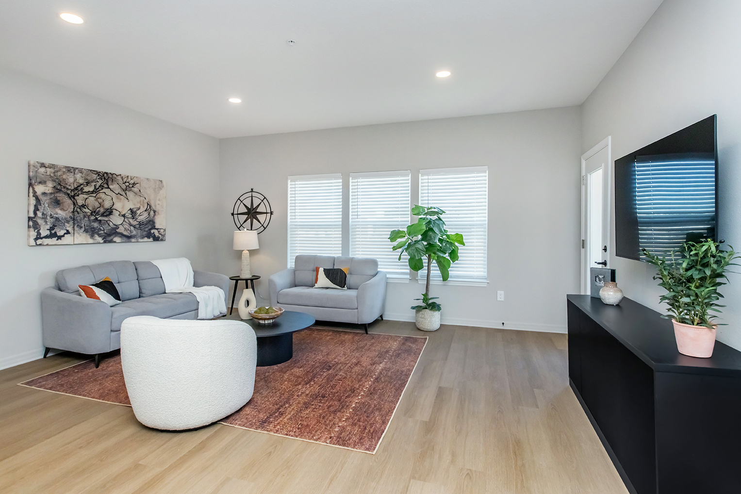 A modern living room featuring two gray sofas with decorative pillows, a round coffee table, and a large potted plant. A wall-mounted TV and a black sideboard are present. Natural light filters through three window blinds, and a colorful area rug adds warmth to the space.
