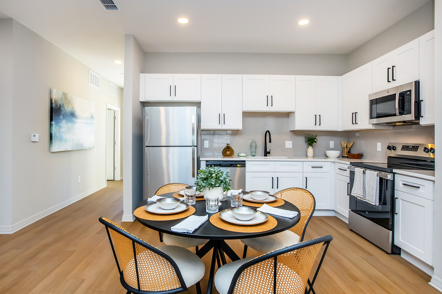 Modern kitchen featuring white cabinets, stainless steel appliances, and a round dining table set for four with stylish plates and glasses. The space has natural light and light wood flooring, creating an inviting atmosphere. A hallway is visible in the background, leading to another area of the home.