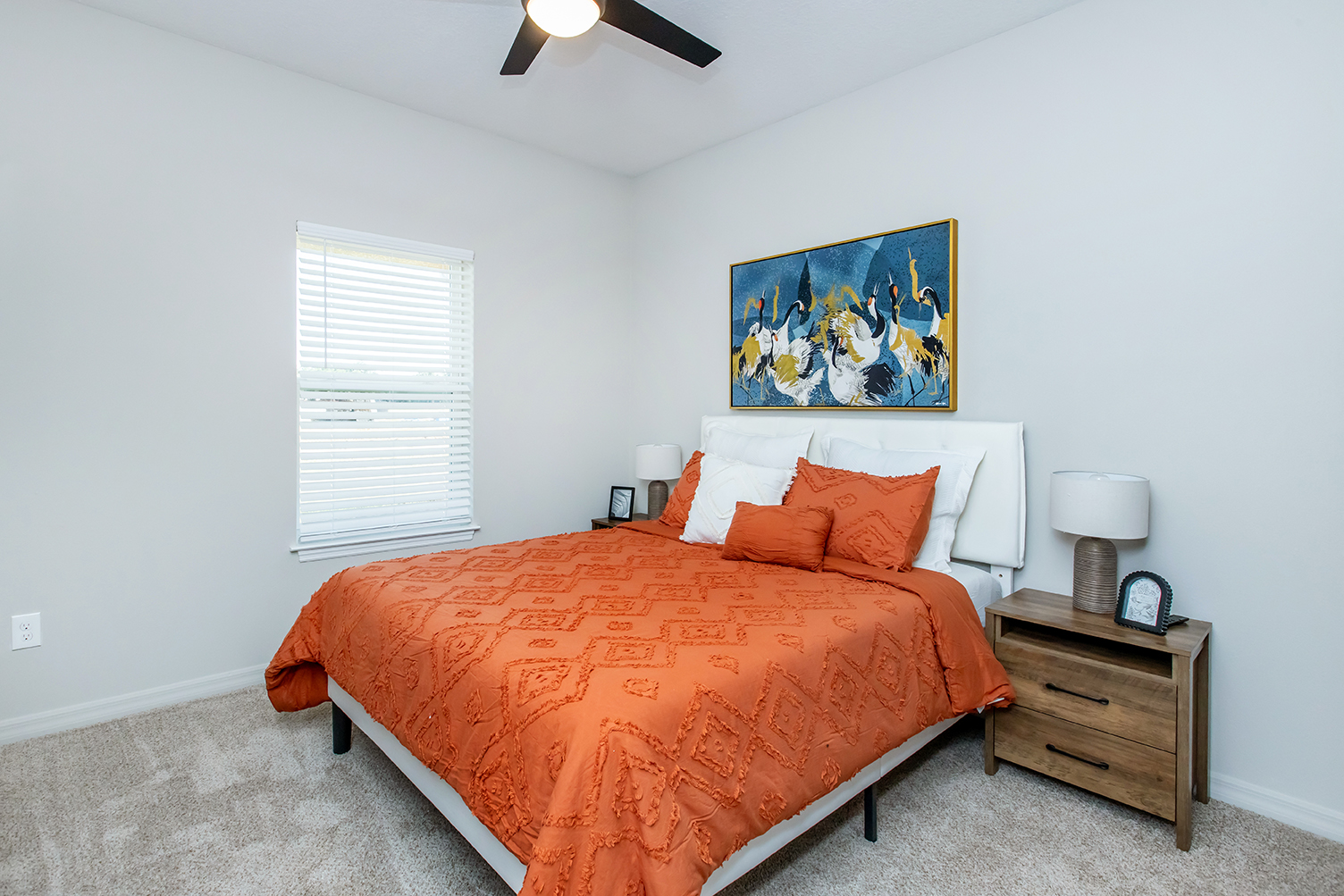 A cozy bedroom featuring an orange bedspread and decorative pillows on a white bed. There's a nightstand with a lamp and clock beside the bed. A colorful painting hangs above the bed, and a window with white blinds allows natural light to fill the room. The walls are painted light, creating a warm atmosphere.