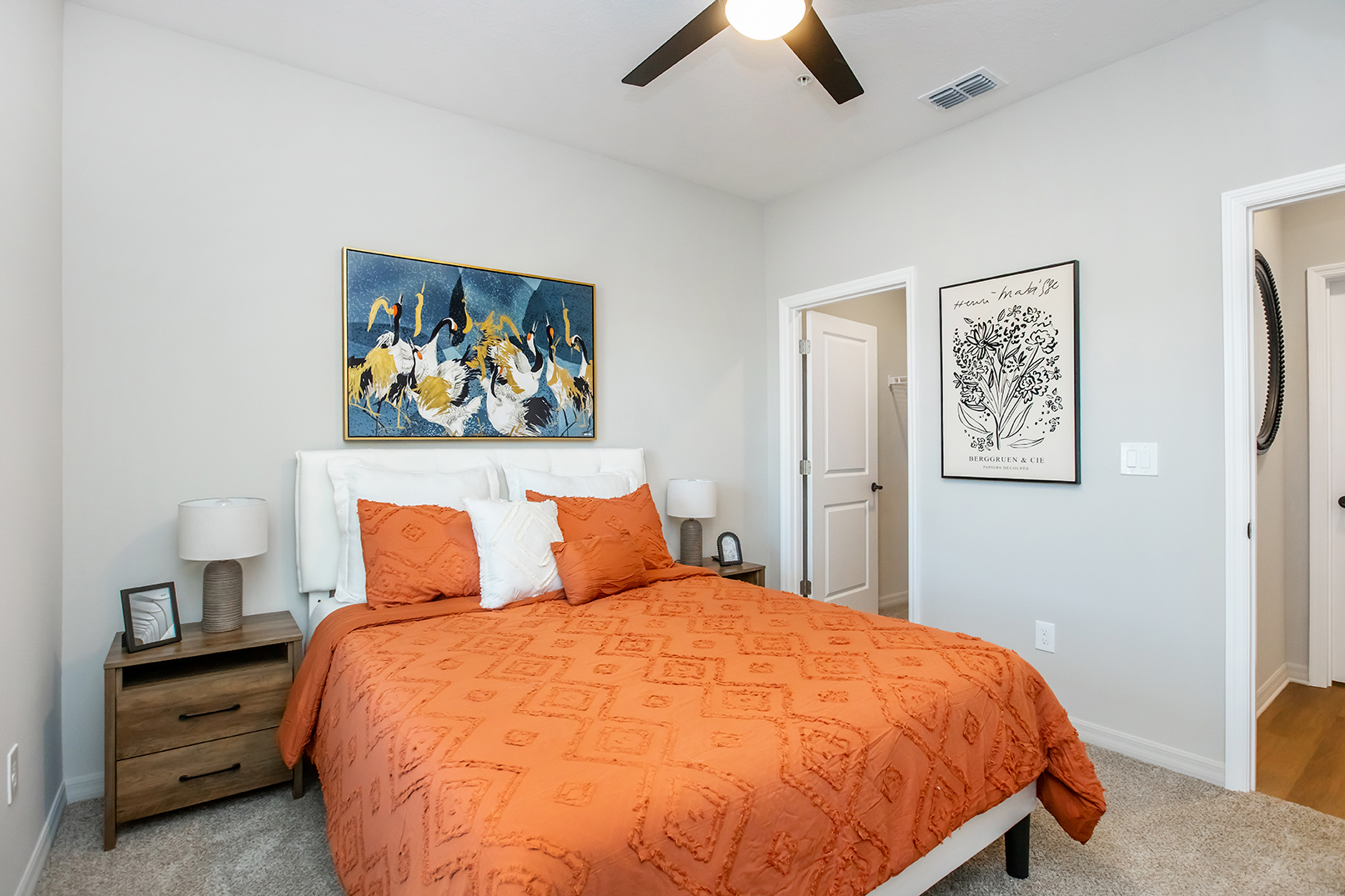 A modern bedroom featuring a queen-sized bed with an orange quilt and decorative pillows. On the wall above the bed, there is a vibrant artwork of birds. The room has light gray walls, a nightstand with a lamp, and a wall art piece on the opposite side. A ceiling fan is also visible.