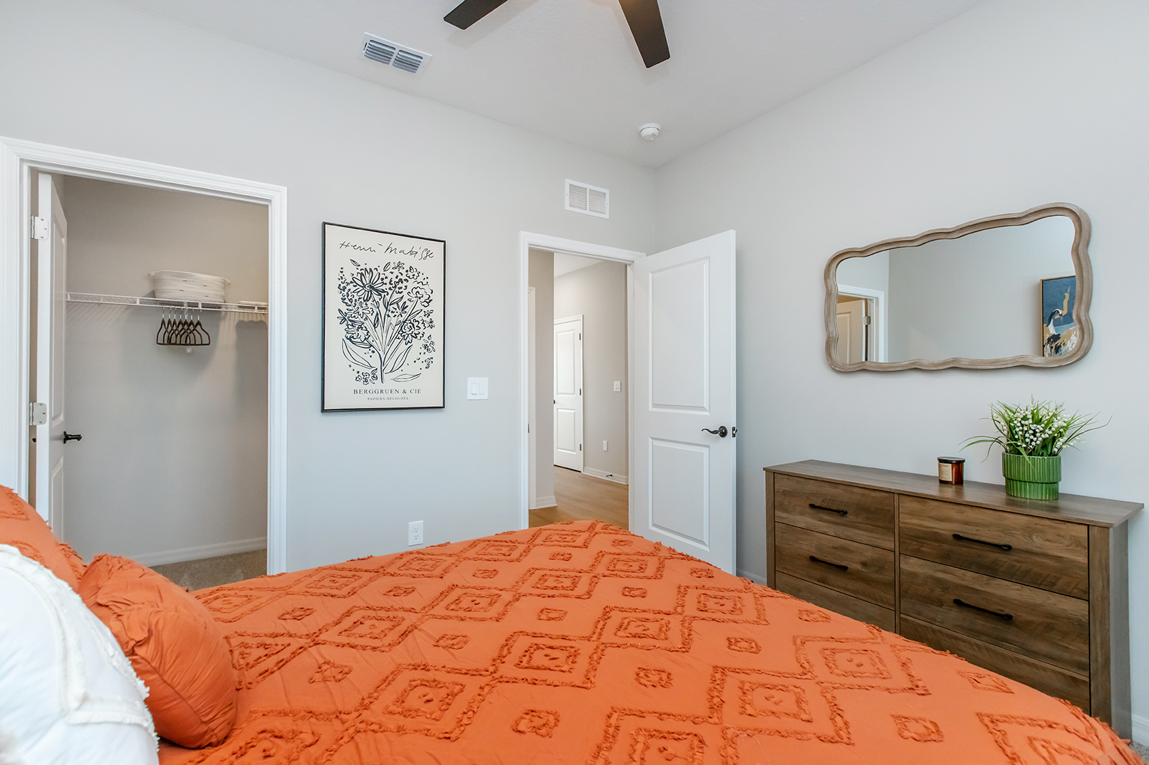 A cozy bedroom featuring an orange patterned bedspread, a wooden dresser, and a mirror on the wall. A closet is visible in the background with shelving. The room has light gray walls and a ceiling fan, creating a welcoming and bright atmosphere.