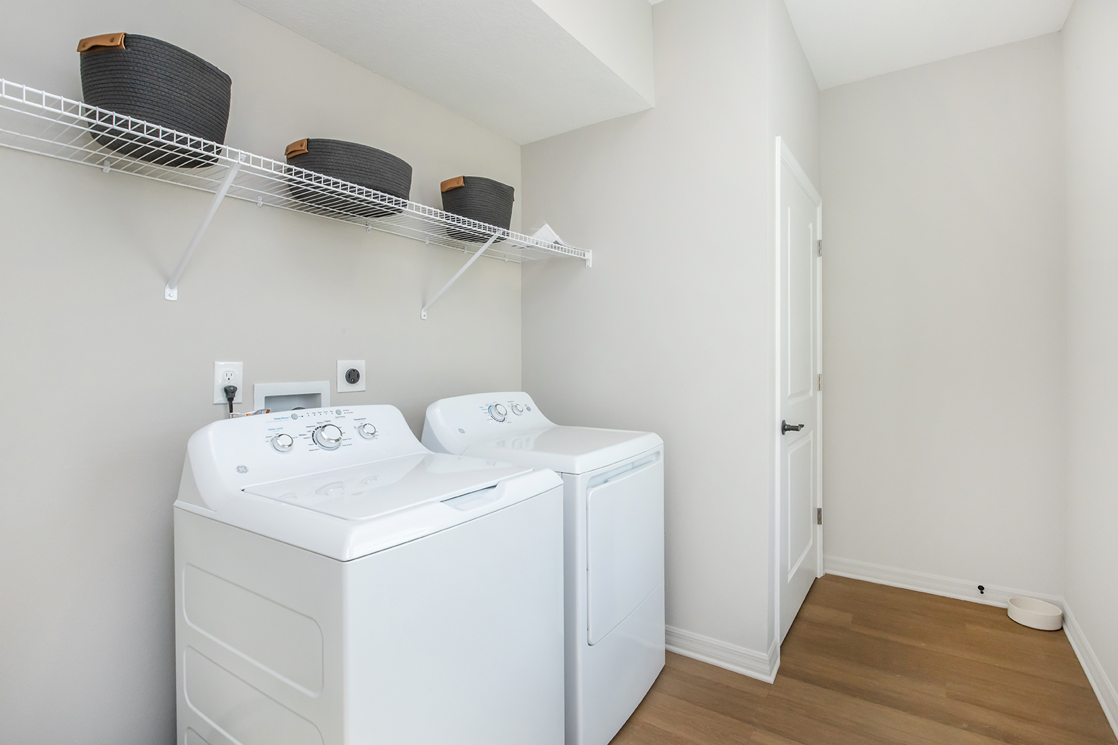 A clean and modern laundry room featuring a white washing machine and dryer side by side. Above them, there are several gray woven baskets on a metal shelf. The walls are painted a light color, and the flooring is a warm wooden finish. A door is visible at the far end of the room.