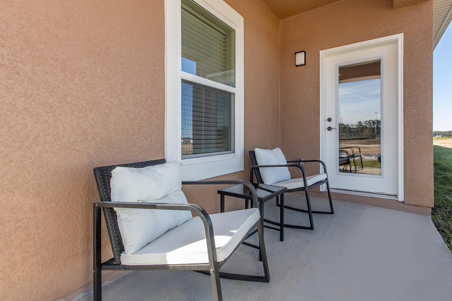 Two outdoor chairs with white cushions are positioned on a porch. The wall is a warm brown color, and there is a glass door reflecting the surrounding area. The porch is well-lit, featuring a window that allows natural light to enter.