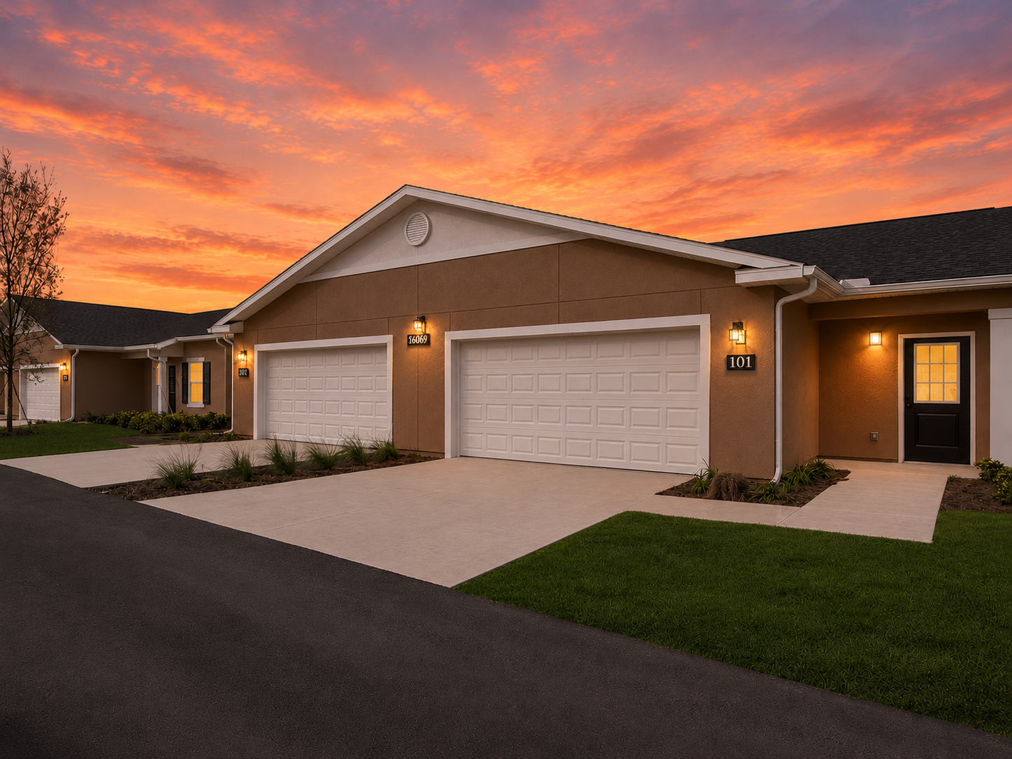 A modern residential building with two garage doors and a welcoming front entrance, set against a vibrant sunset sky. The surroundings are landscaped with grass and plants, contributing to a tranquil atmosphere in a suburban neighborhood.
