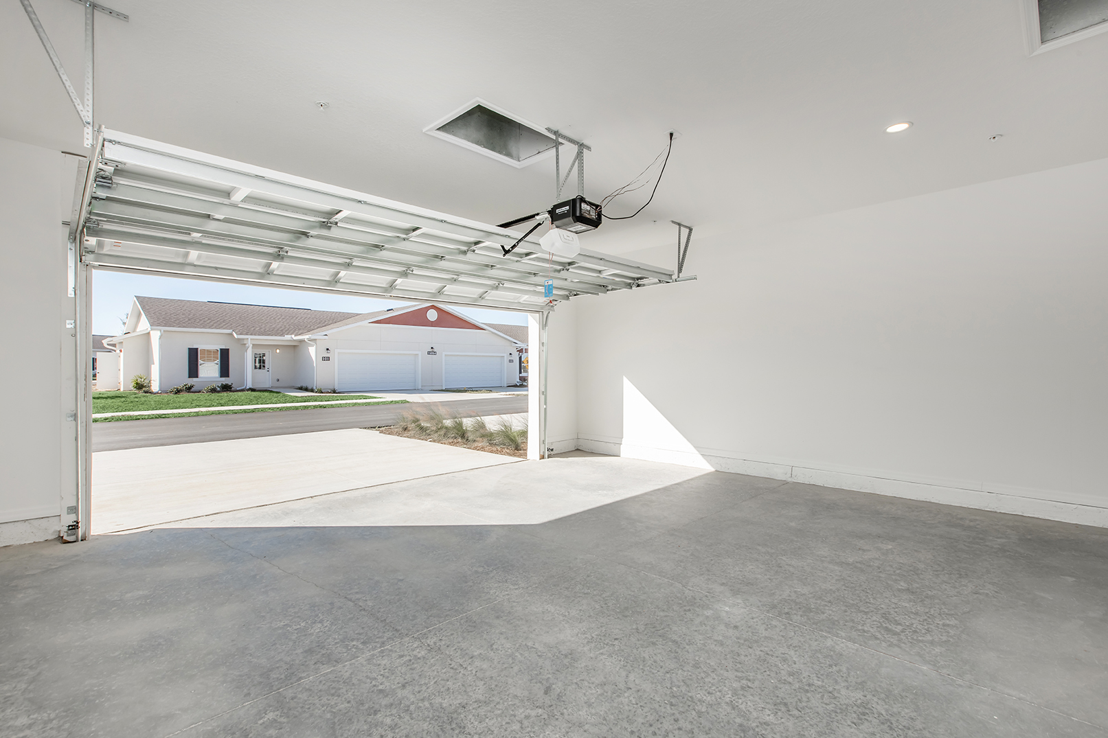 A spacious, clean garage with an open door, revealing a suburban street outside. The garage has a concrete floor and white walls, illuminated by natural light. In the background, there are houses visible across the street.