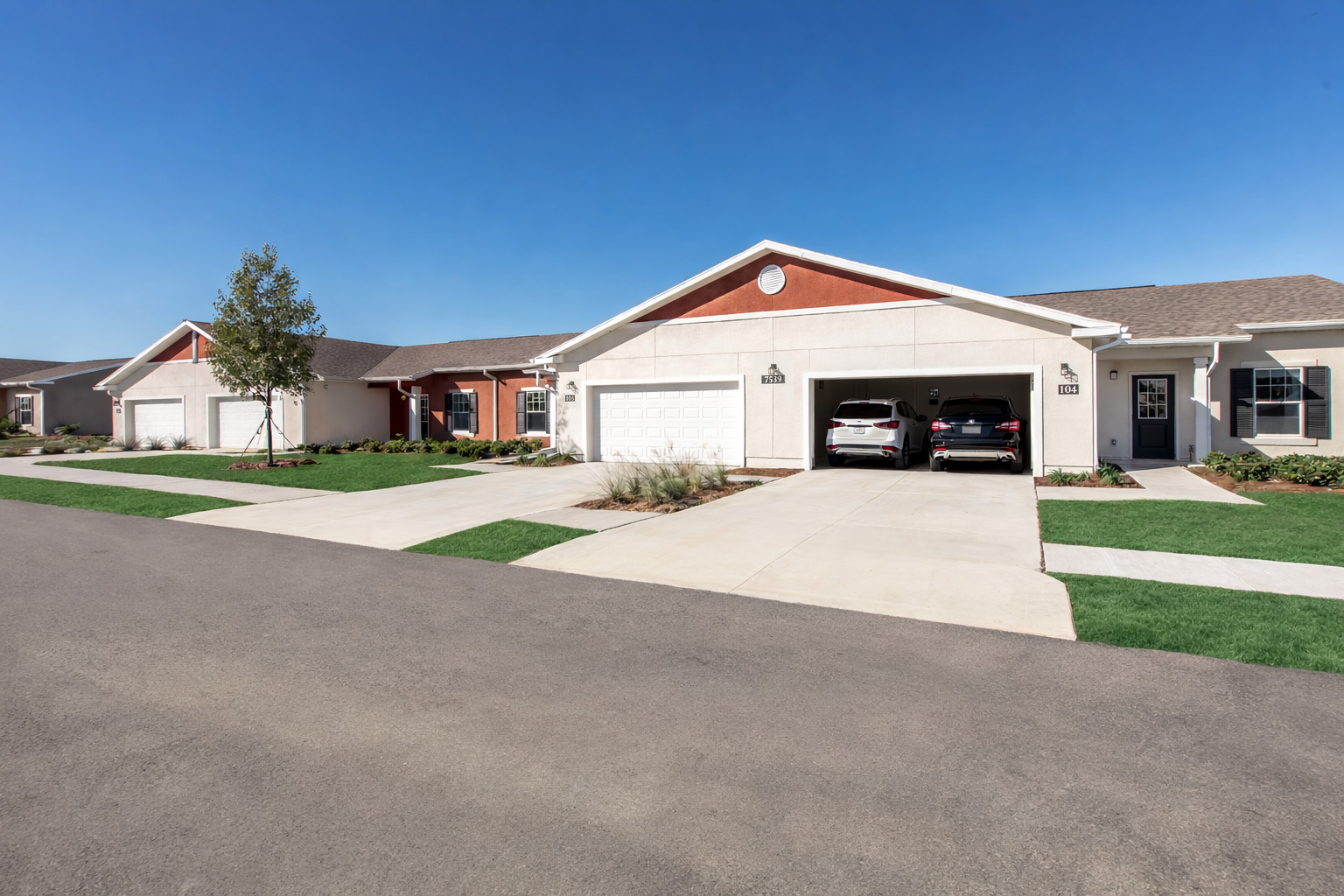 A residential neighborhood featuring single-story homes with attached garages. The houses have neat landscaping, a clear blue sky overhead, and a well-maintained street without visible traffic. Two cars are parked in front of one of the garages, and the area appears spacious and inviting.